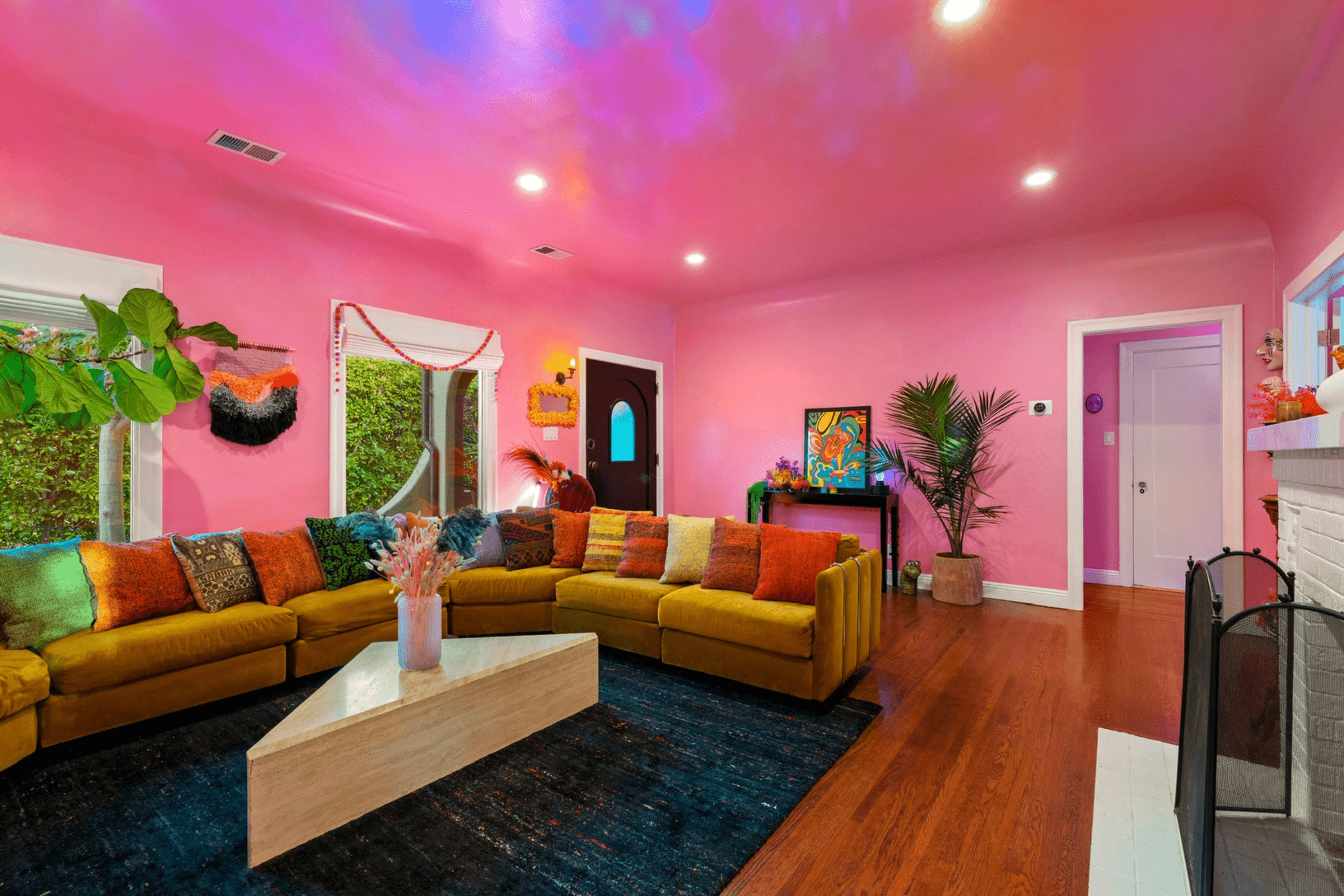 A bright pink living room features a large, curved yellow sectional sofa, a geometric coffee table, and a vibrant wall decor.