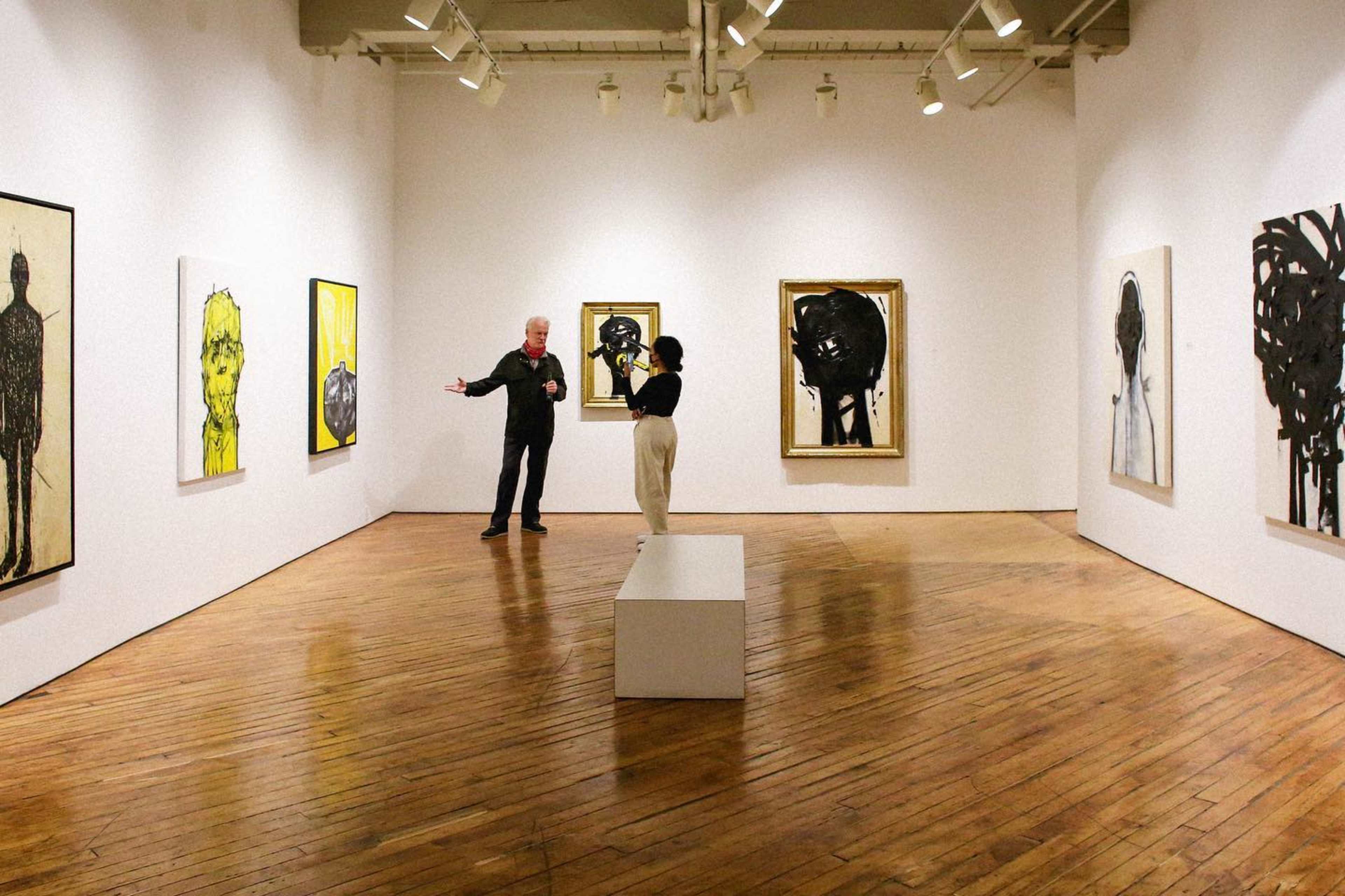A man and a woman are discussing art in a gallery filled with large paintings on white walls and a wooden floor.