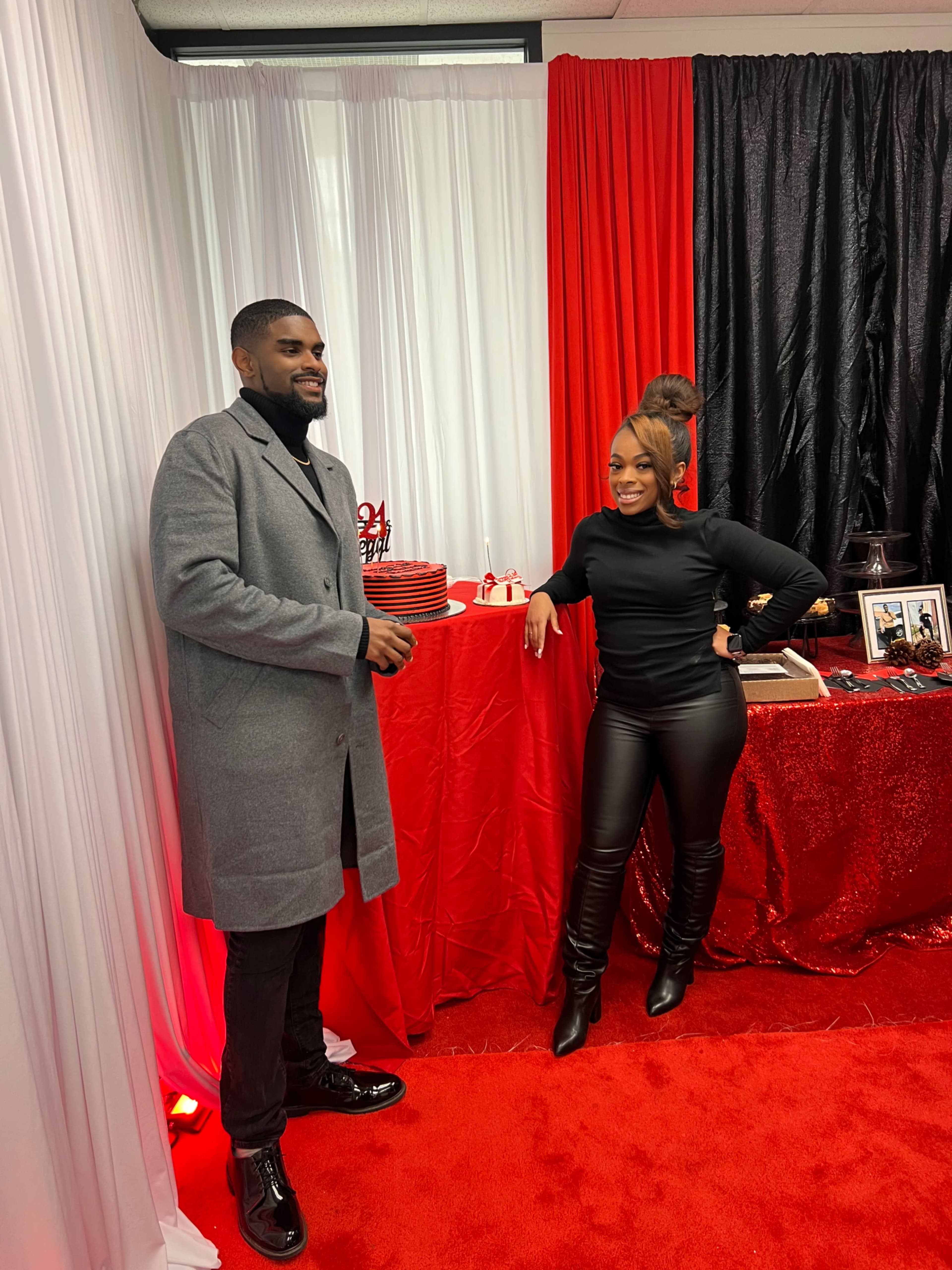 A man in a gray coat stands next to a woman in a black top and leather pants in front of a decorated table featuring a cake and red accents.