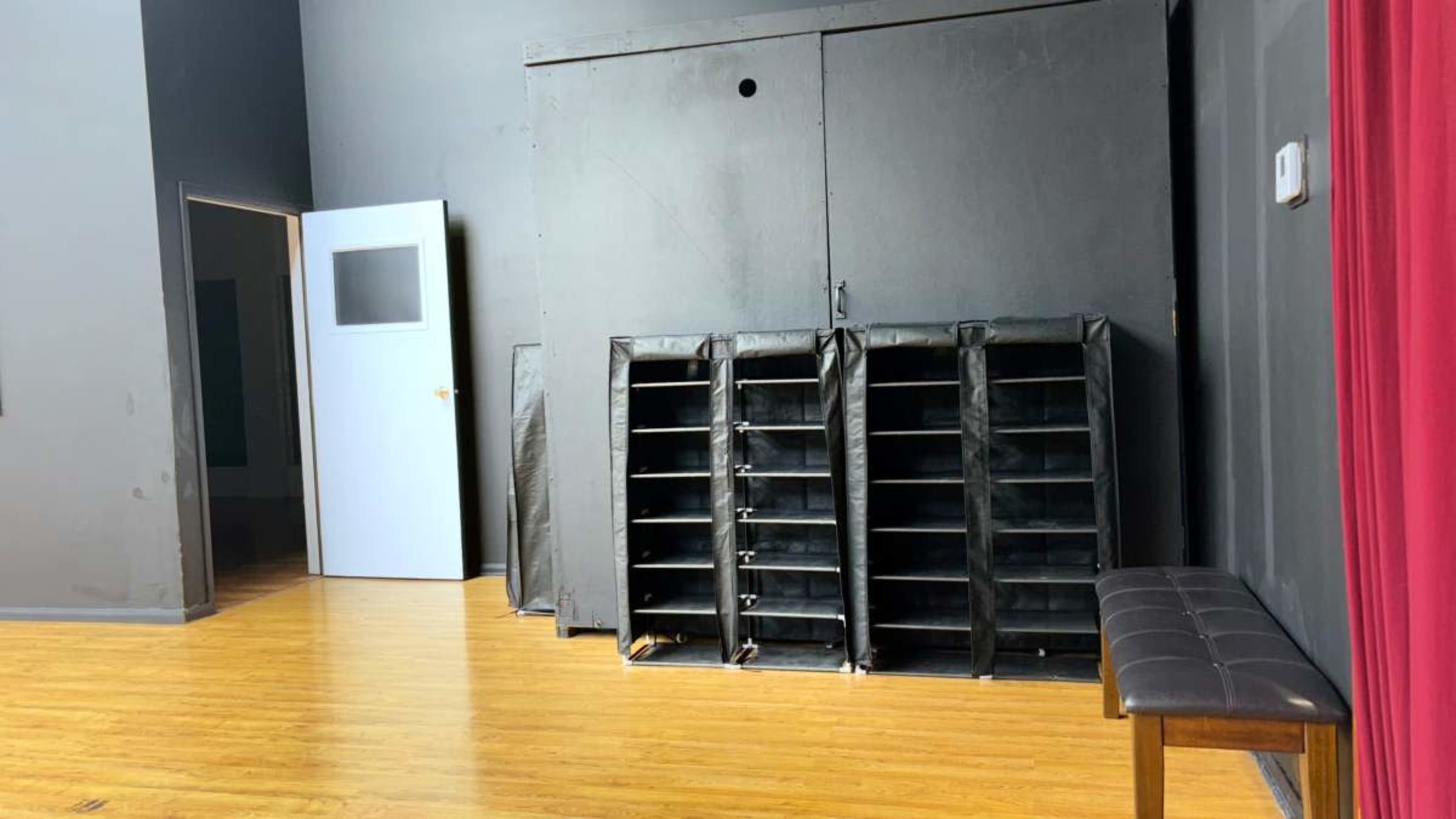 The image shows a room with dark walls, a closed door on the left, a large black storage unit against the wall, and a brown bench in front of it.