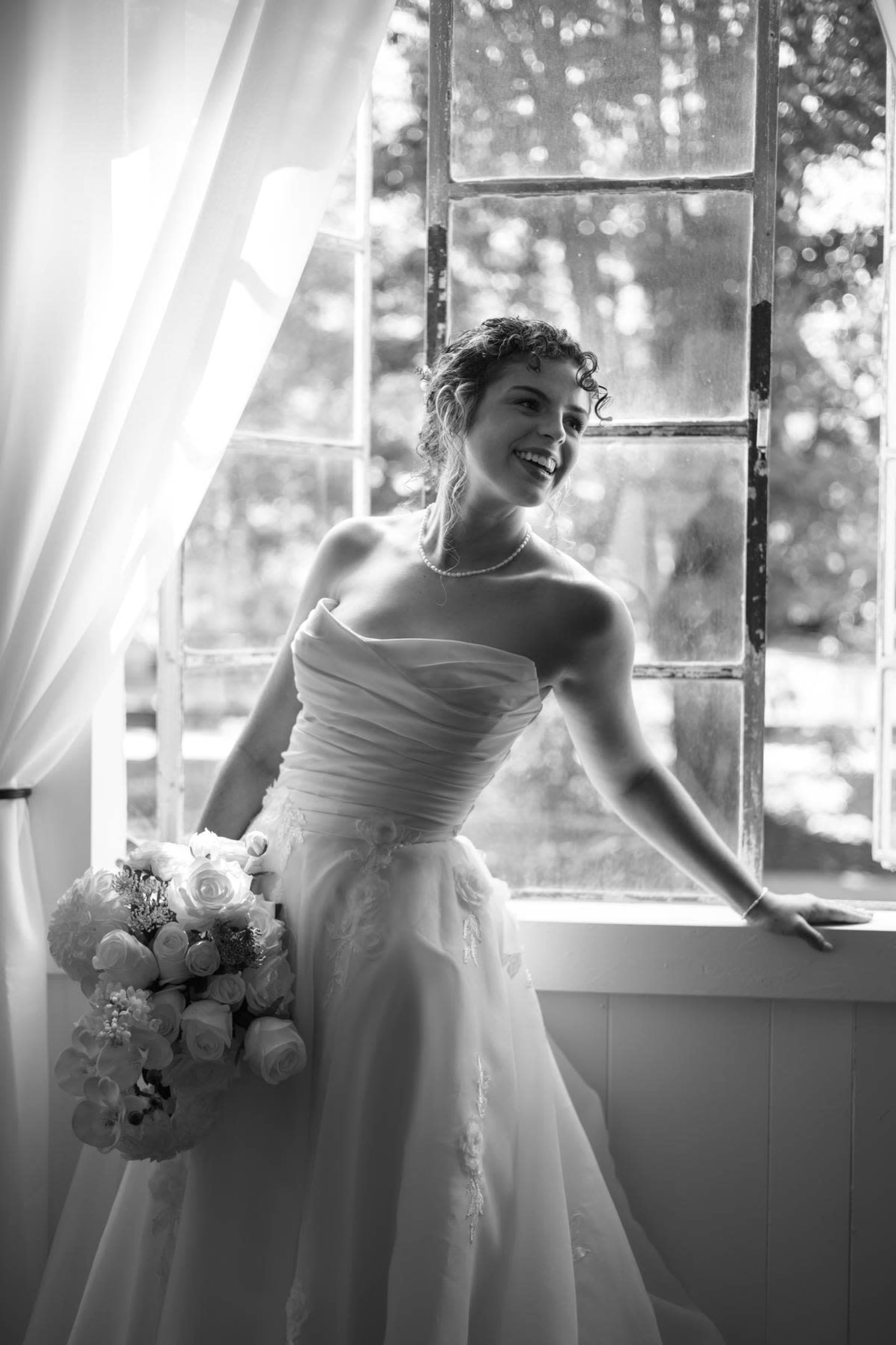 A bride in a strapless white dress stands by an open window, holding a bouquet of flowers and smiling while looking outside.