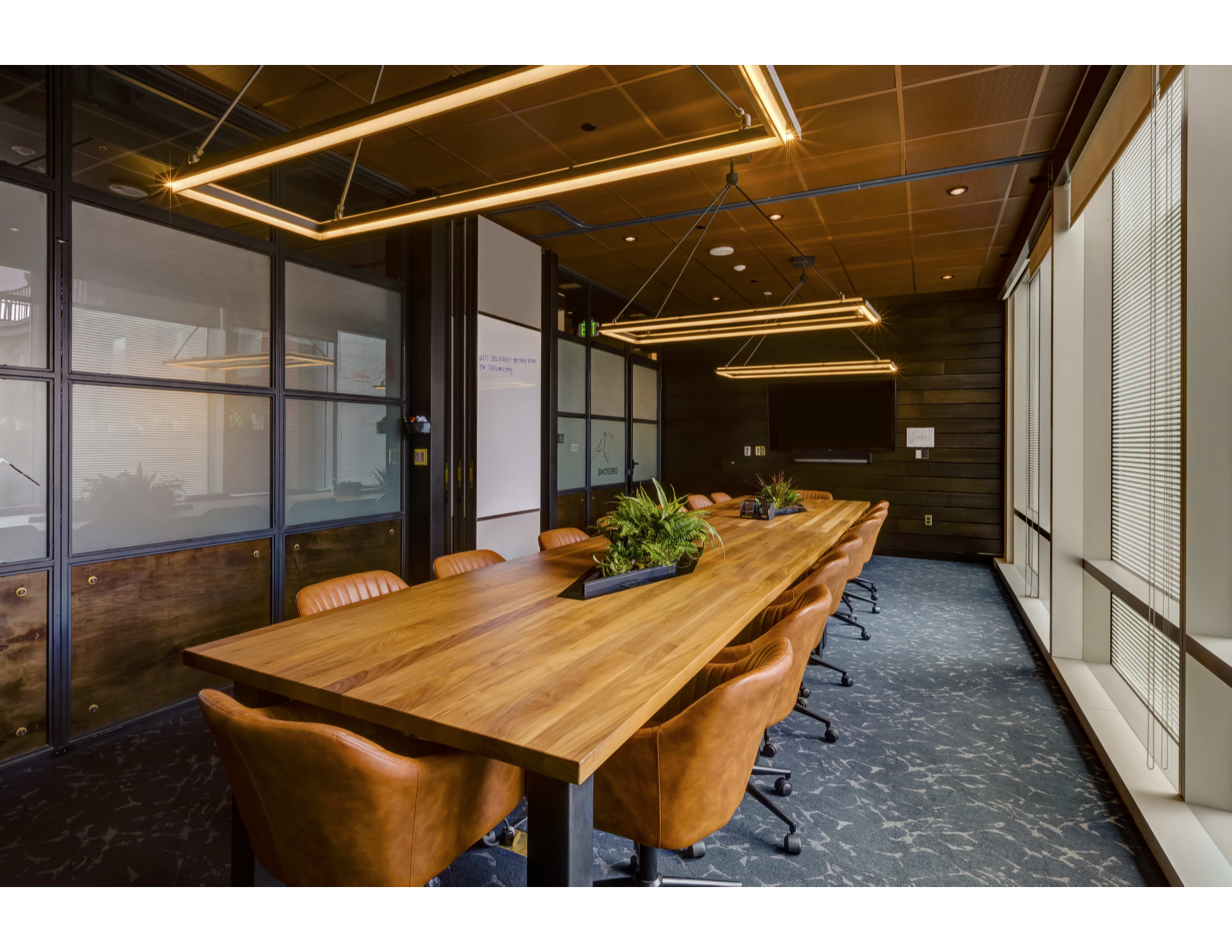 A long wooden conference table with leather chairs is placed in a modern office meeting room featuring large windows and suspended light fixtures.