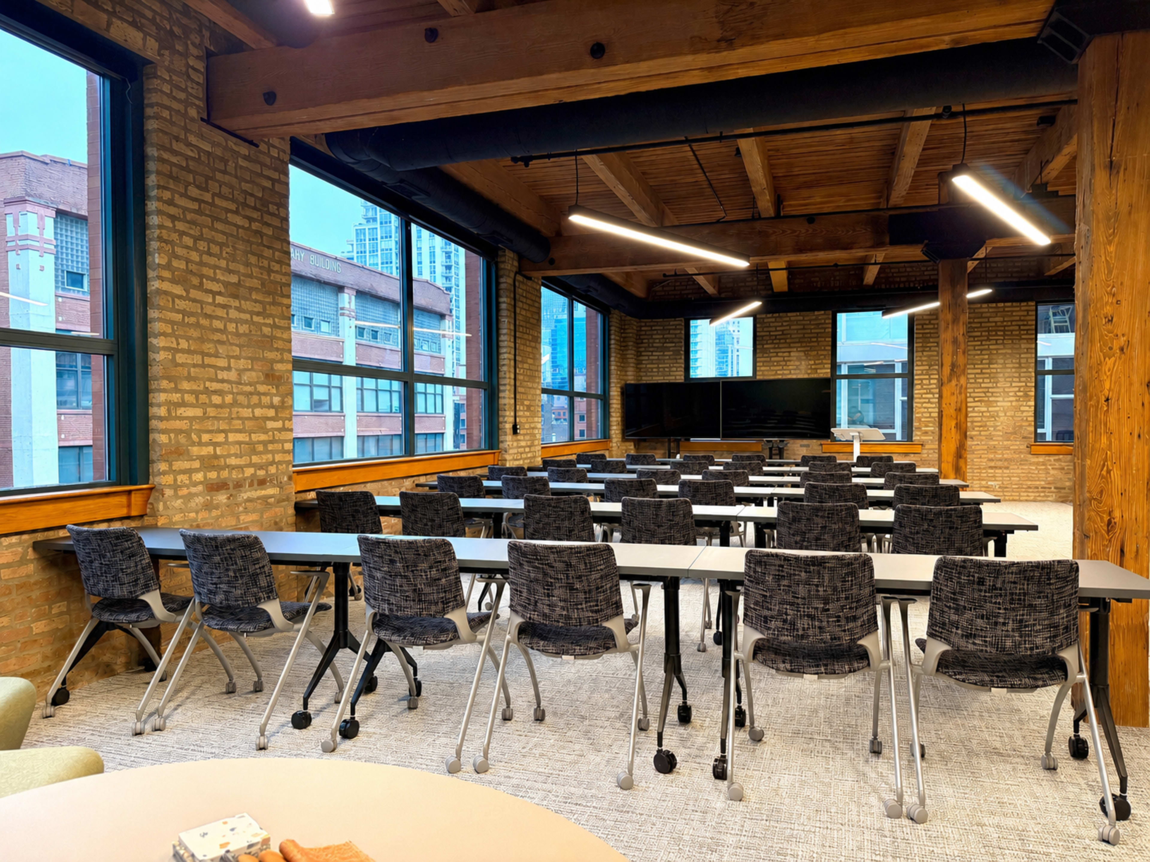 The image shows a classroom setting with rows of grey tables and black woven chairs, featuring large windows that let in natural light.