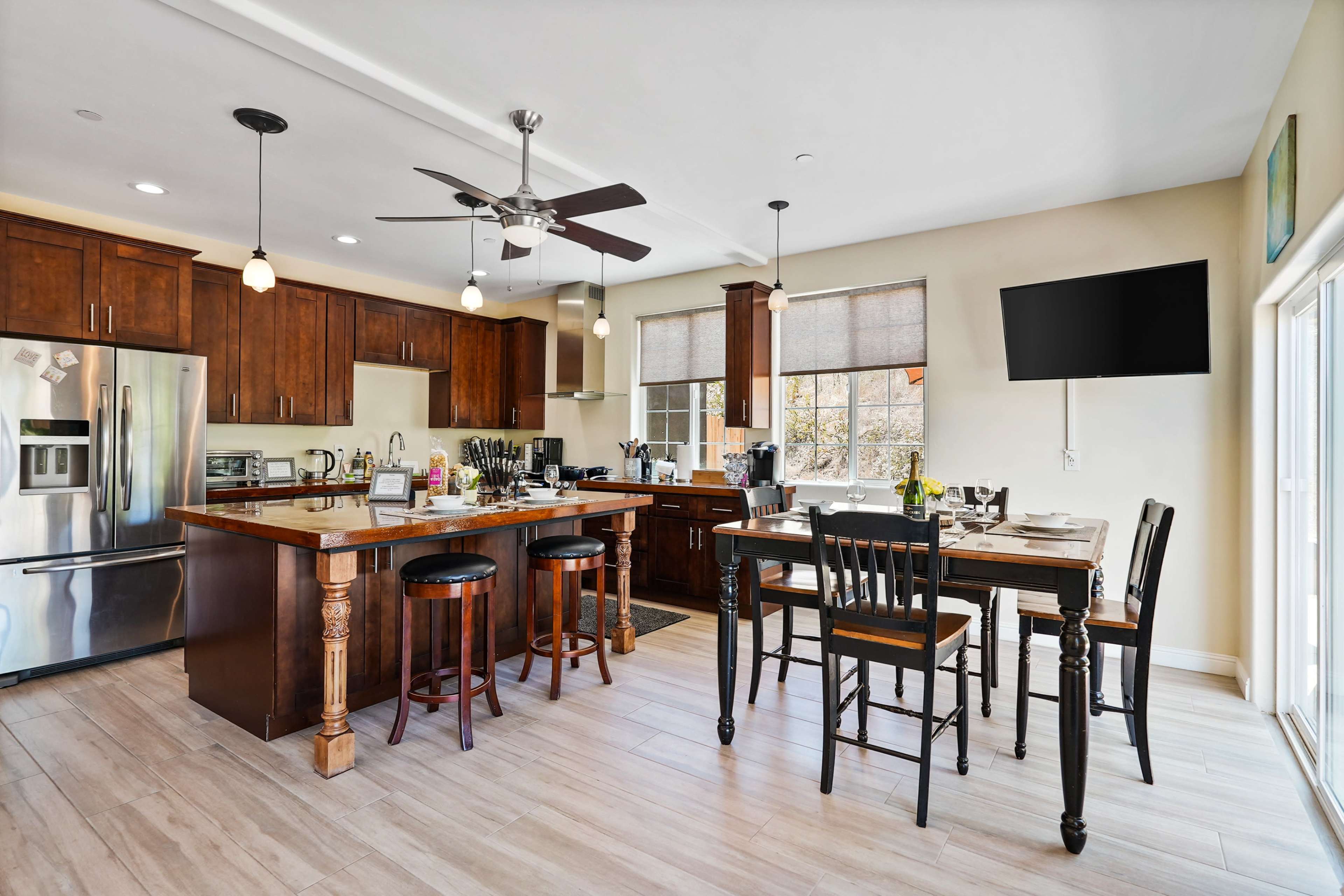 A modern kitchen features wooden cabinets, a central island with bar stools, and a dining table in front of large windows.