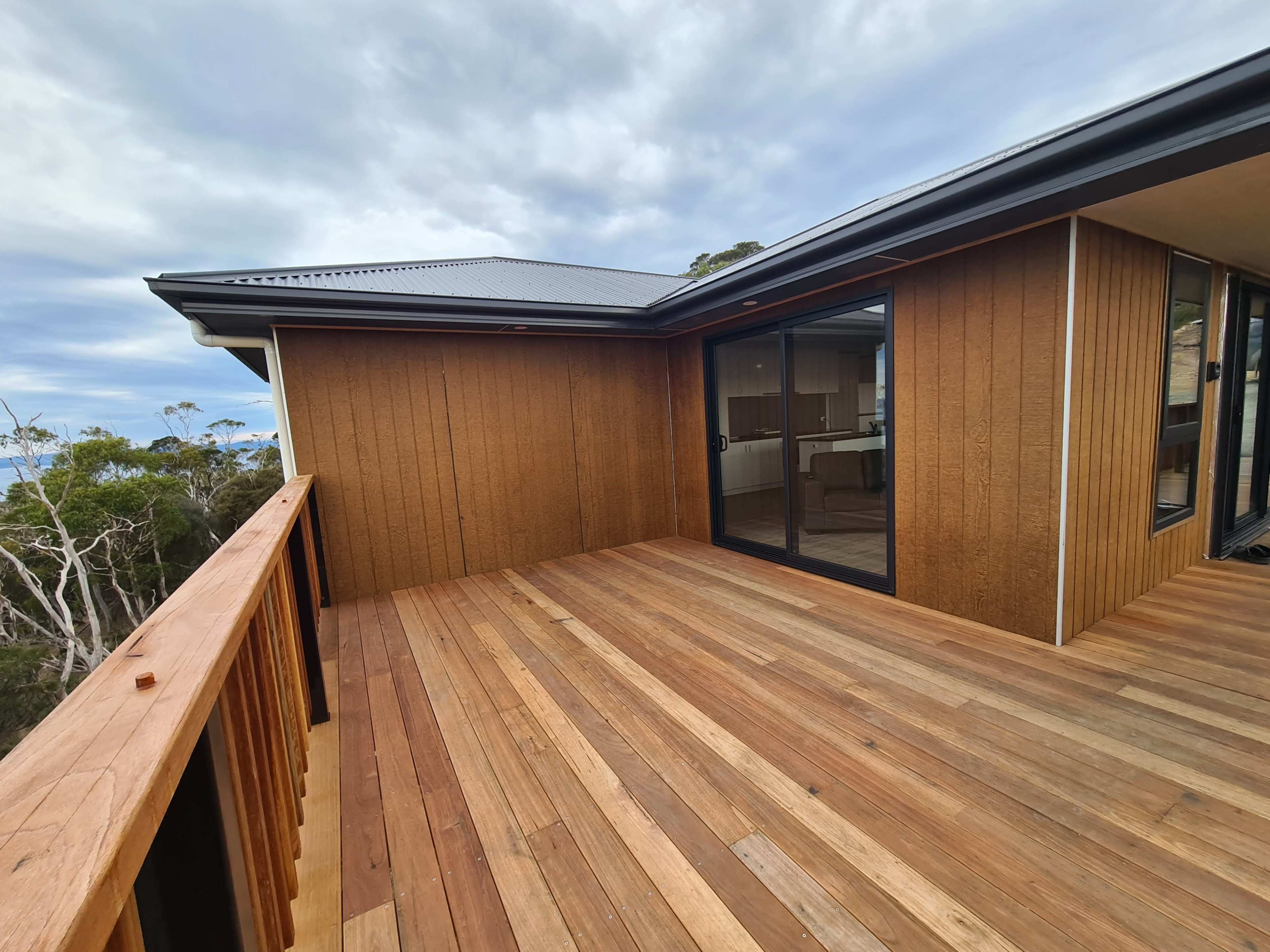 A wooden deck extending from a house, with a view of trees and cloudy sky in the background.