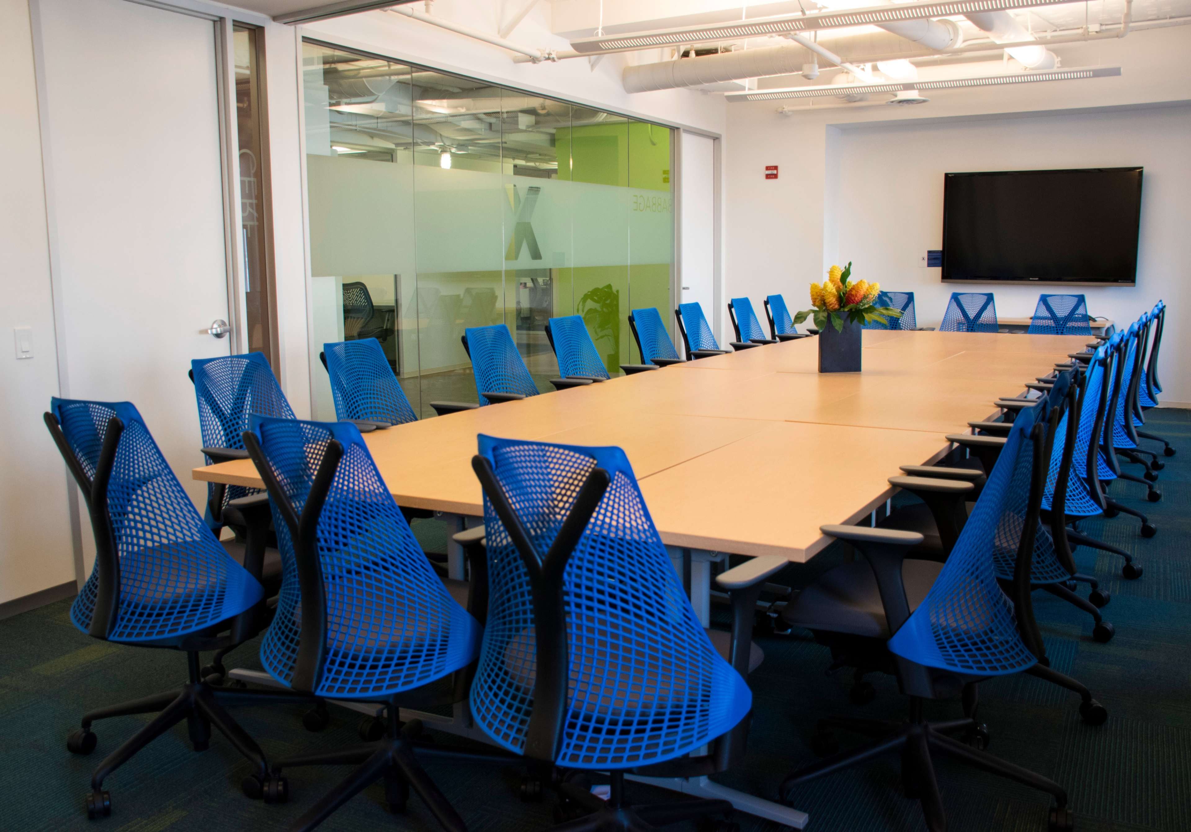 A large conference room features a long table surrounded by blue mesh office chairs, with a television screen mounted on the wall and a glass wall displaying an office space.