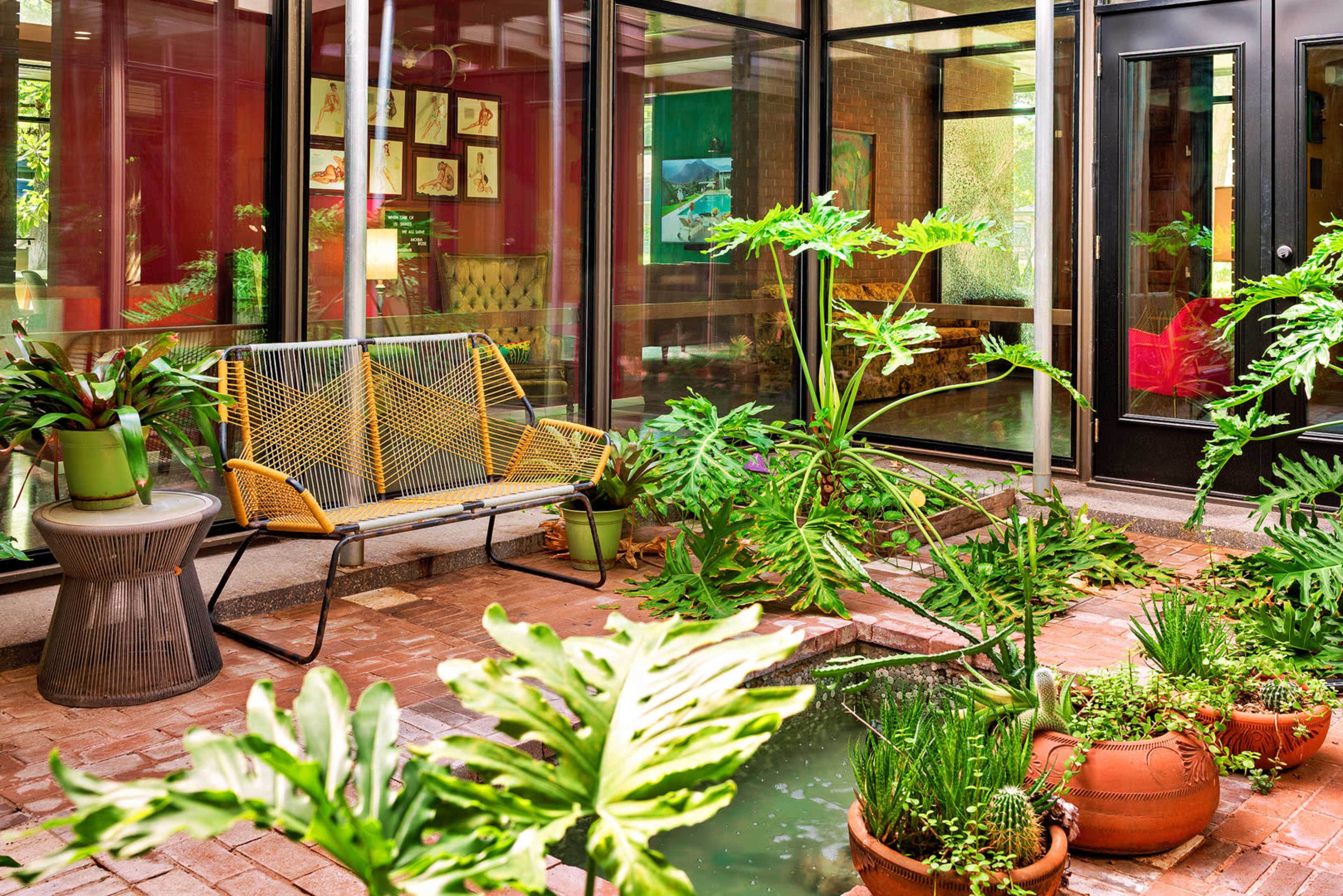 The image shows a vibrant indoor garden with potted plants, a small water feature, and a yellow seating area surrounded by glass walls.