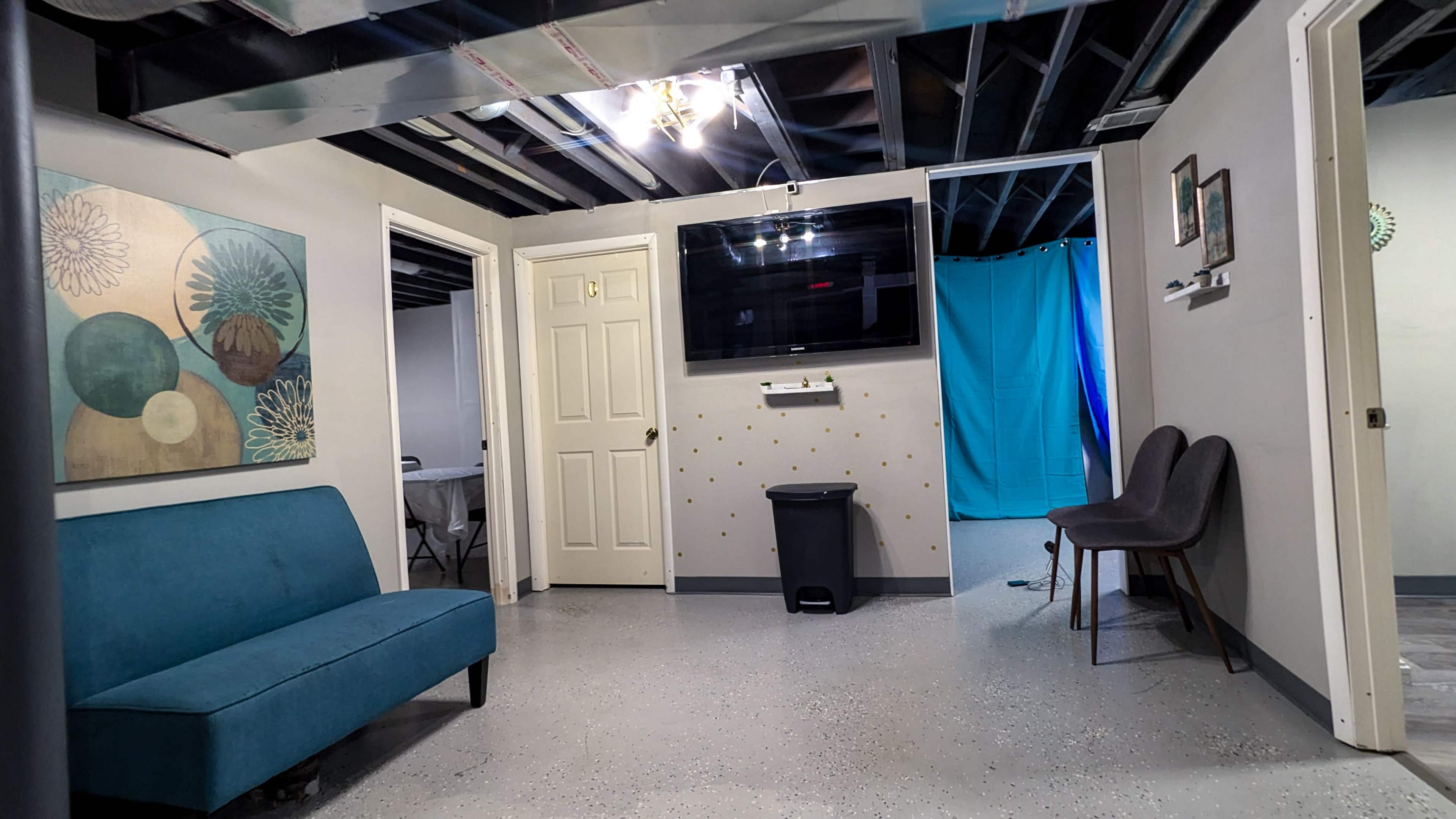The image shows a basement room with a blue couch, a wall-mounted television, and two chairs near a door leading to a bathroom or another room, with a blue curtain visible in the background.