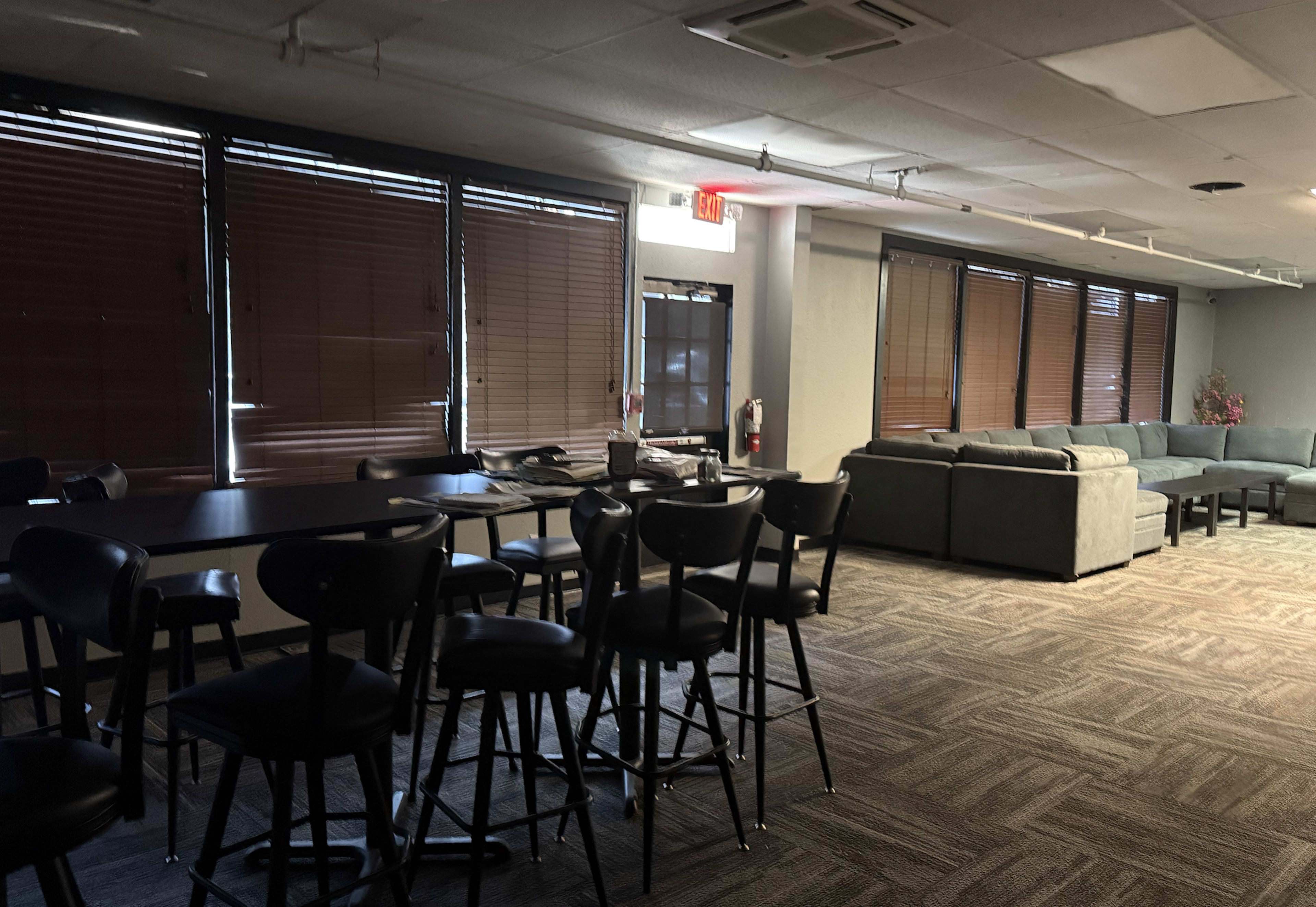 The image shows a room with a seating area that includes a gray couch, a table with chairs, and large windows covered by wooden blinds.