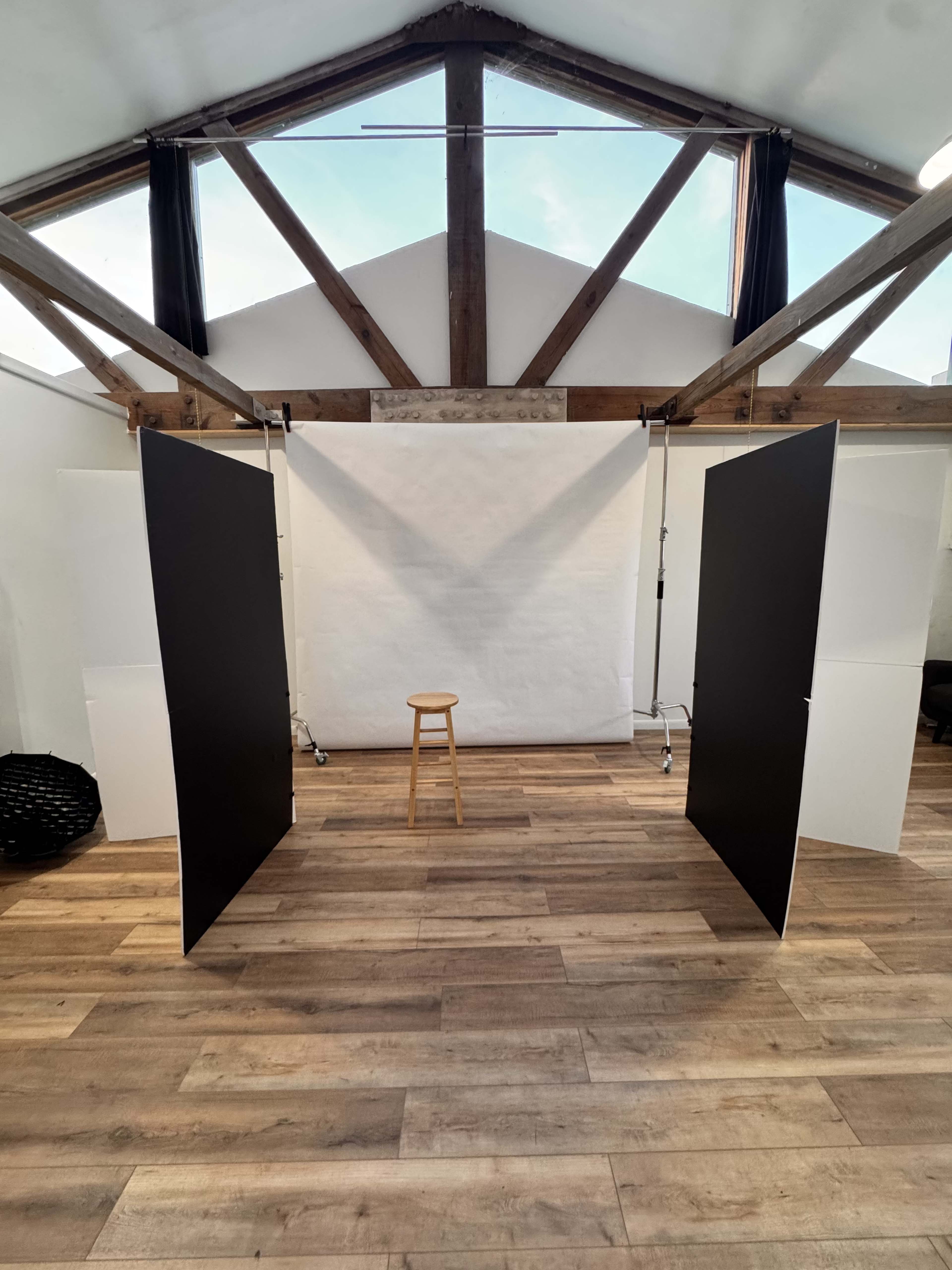 The image shows a photography studio featuring a wooden floor, a stool in the center, and two large black panels positioned on either side of a white backdrop.