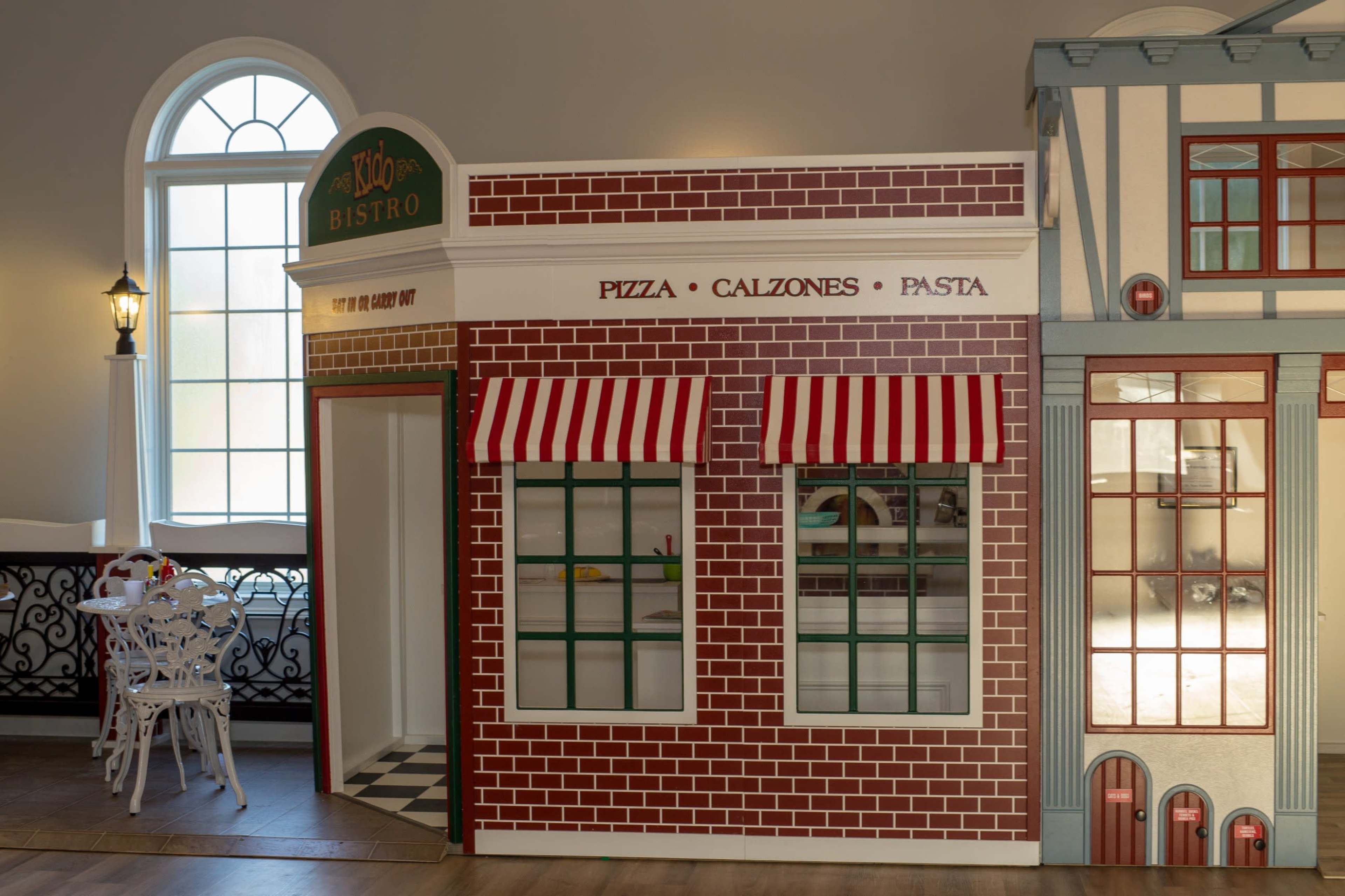 The image shows a colorful play restaurant facade with a brick exterior and striped awnings, featuring signs for pizza, calzones, and pasta.