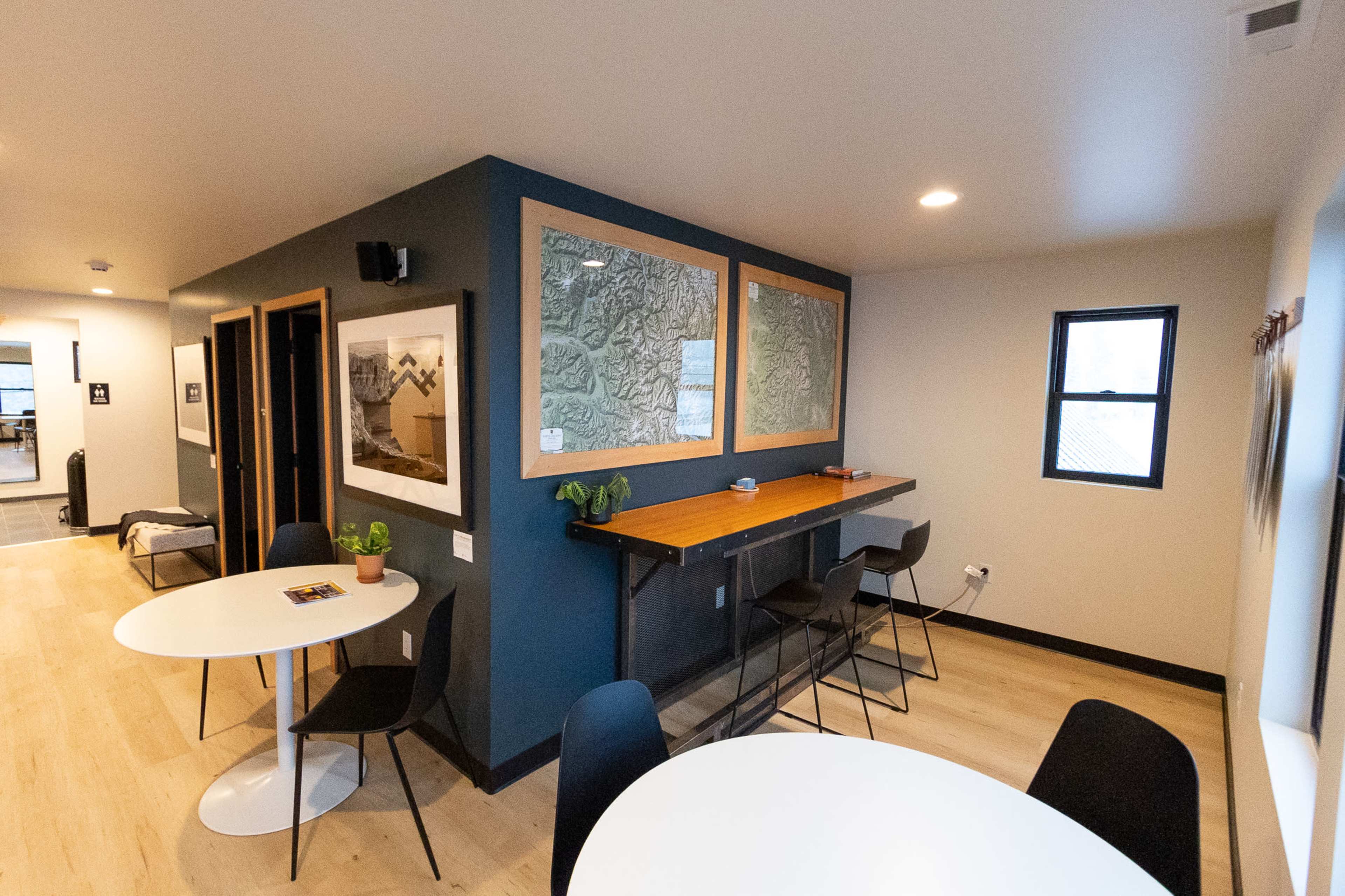 The image shows a modern interior space featuring a bar-height table with stools, two round white tables, and a combination of framed art and textured glass panels on the walls.
