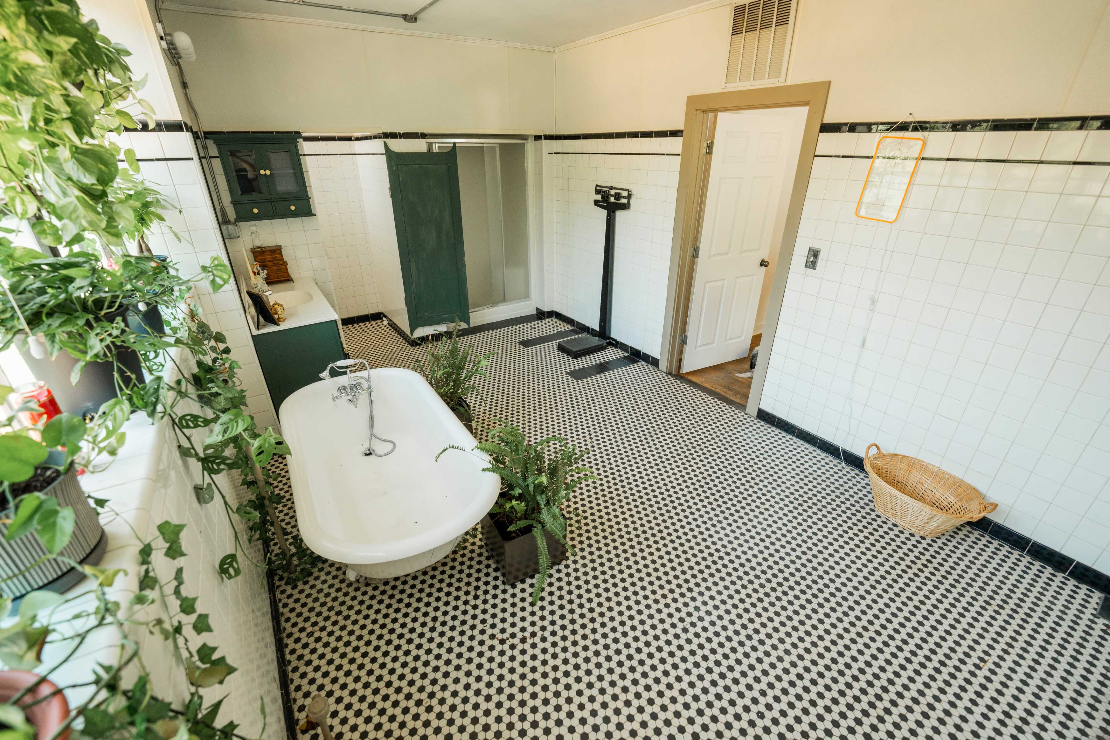 A bright bathroom with black-and-white tiled flooring, a vintage clawfoot bathtub, a scale, and potted plants against the walls.
