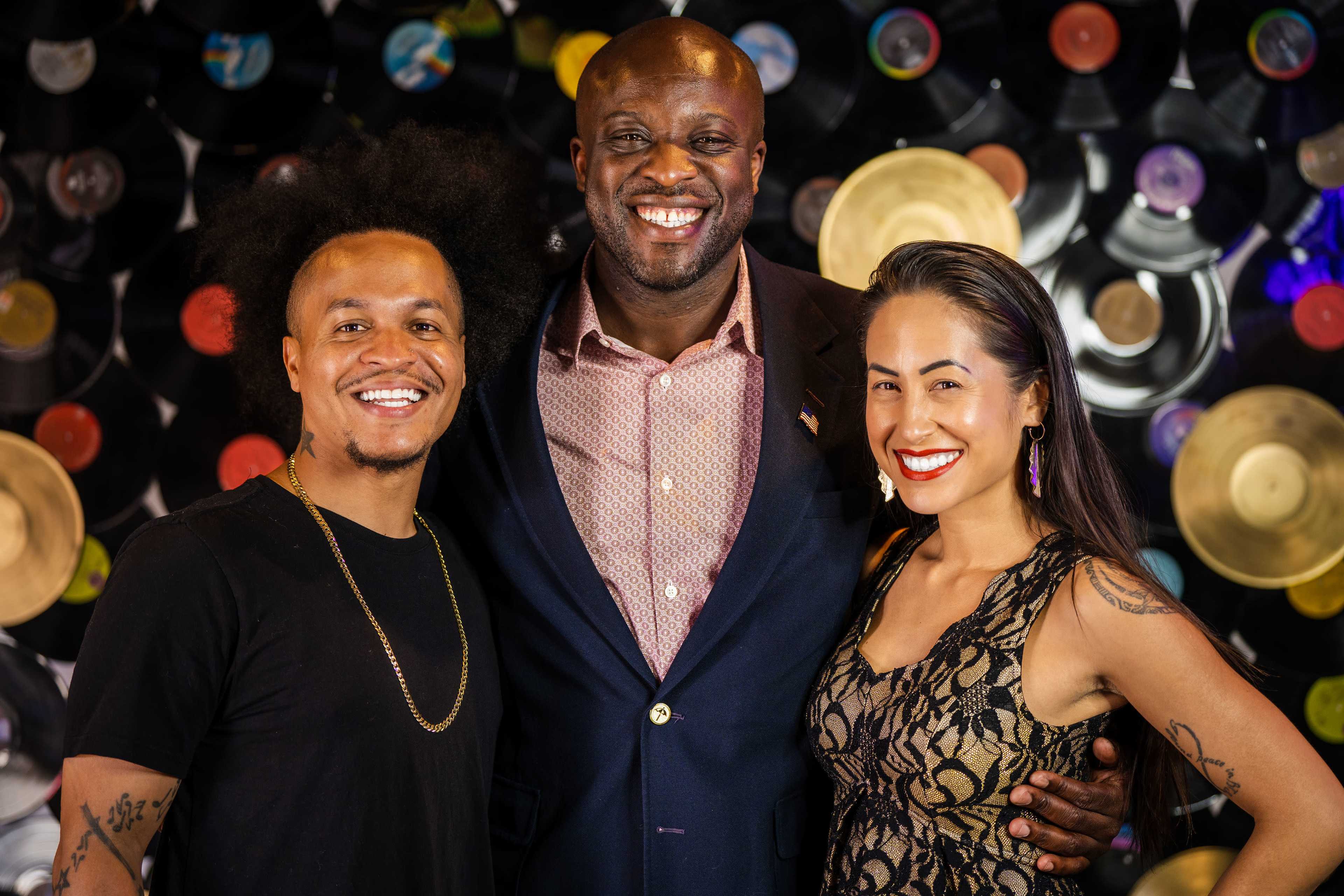 Three individuals pose for a photo in front of a decorative backdrop made of vinyl records.