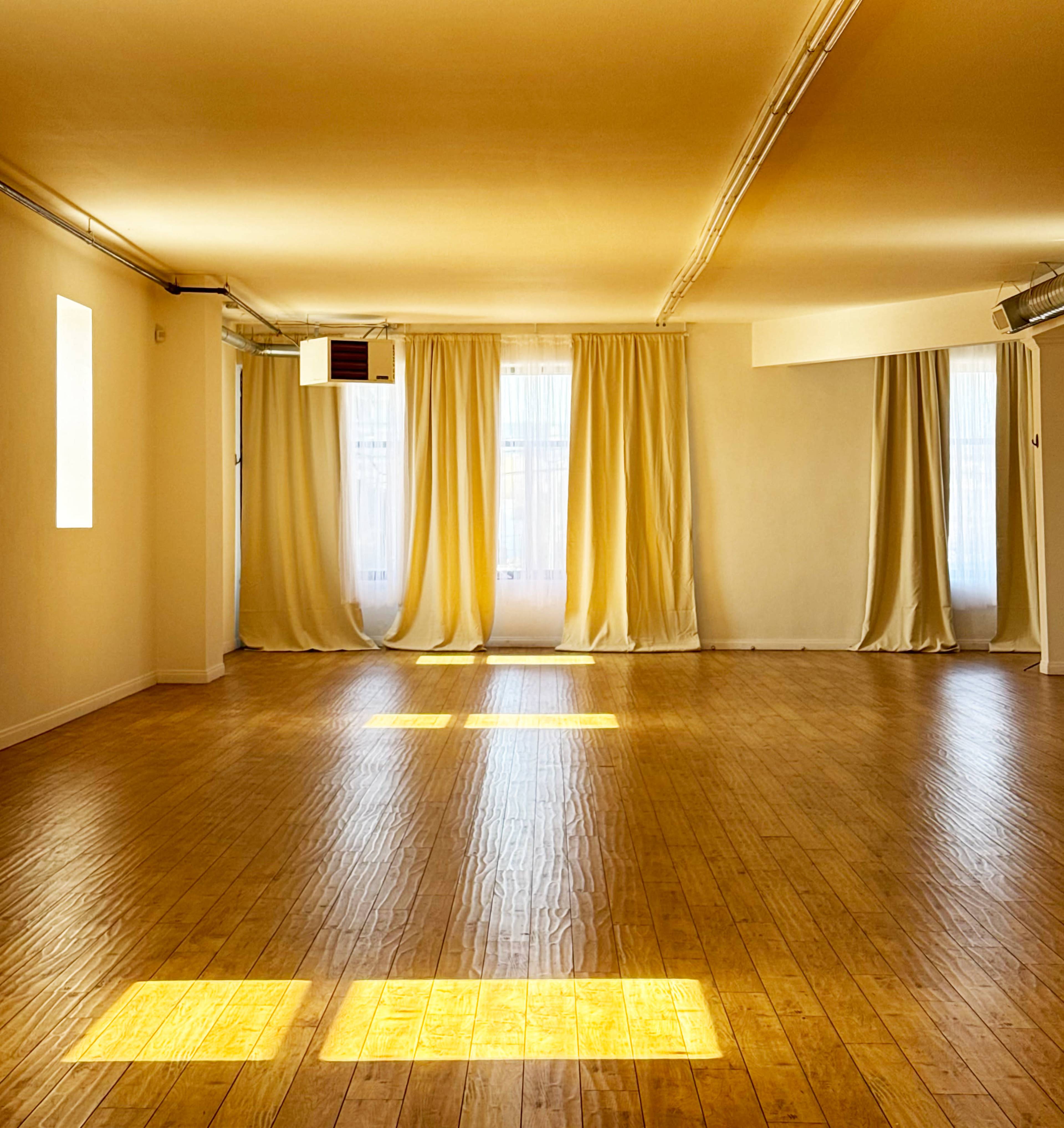 The image shows a spacious, brightly lit room with wooden flooring, large windows covered by sheer curtains, and minimal decoration.