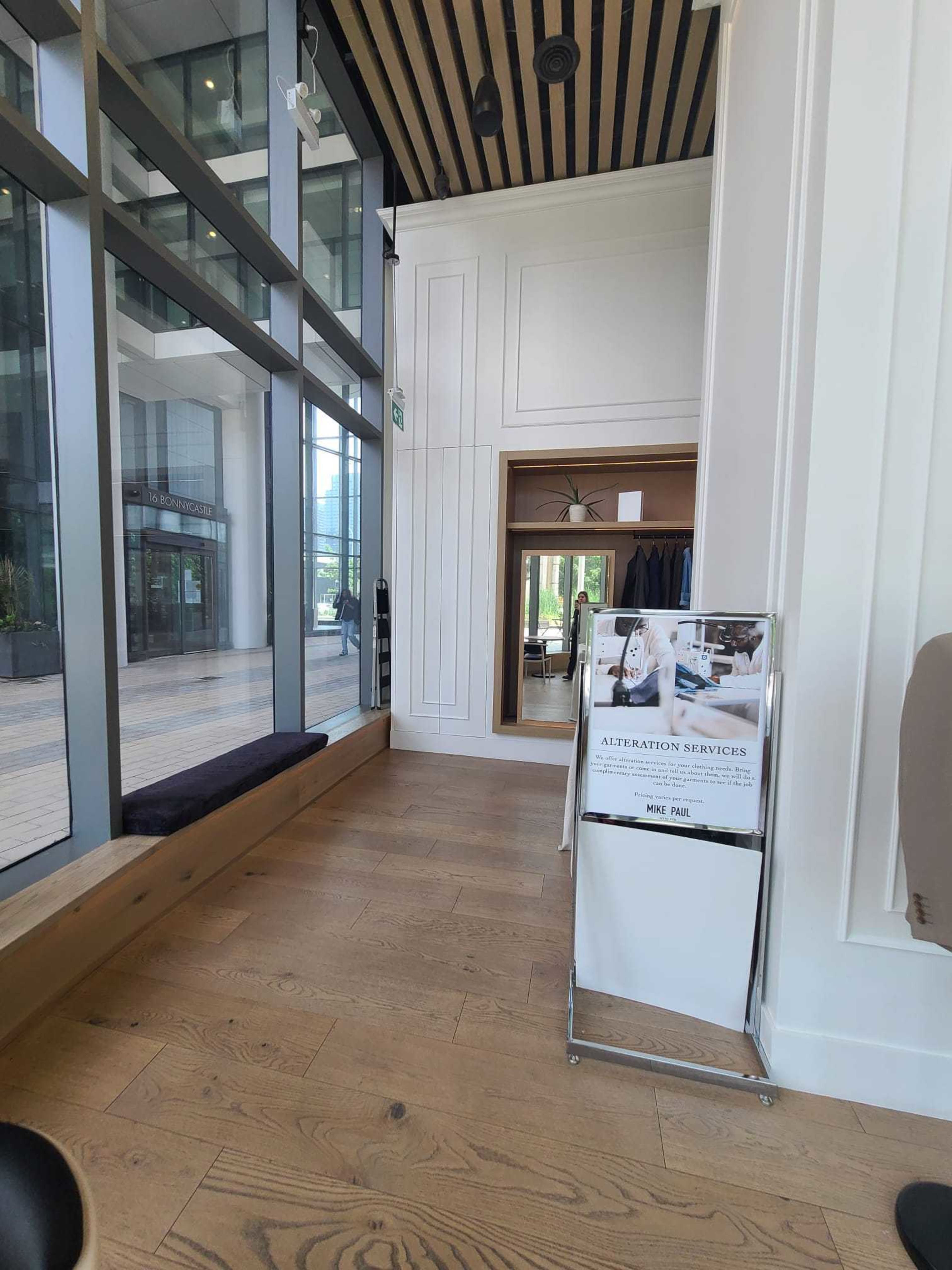 The image shows a well-lit interior space featuring a glass wall, wooden flooring, and a sign for alteration services near a mirror.