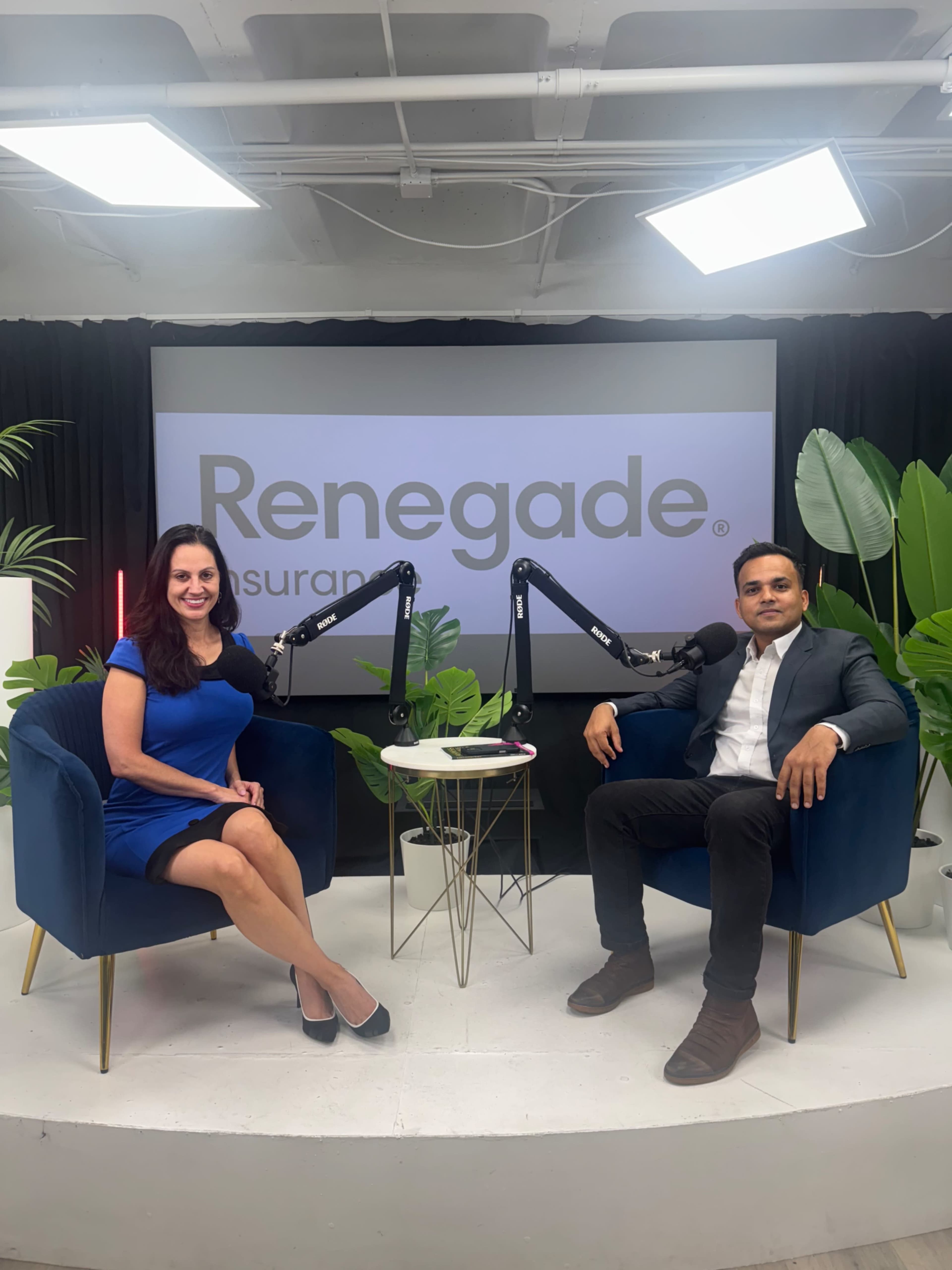 A woman in a blue dress and a man in a suit sit in chairs facing the camera in a well-lit studio setting featuring a large screen displaying the "Renegade Insurance" logo.