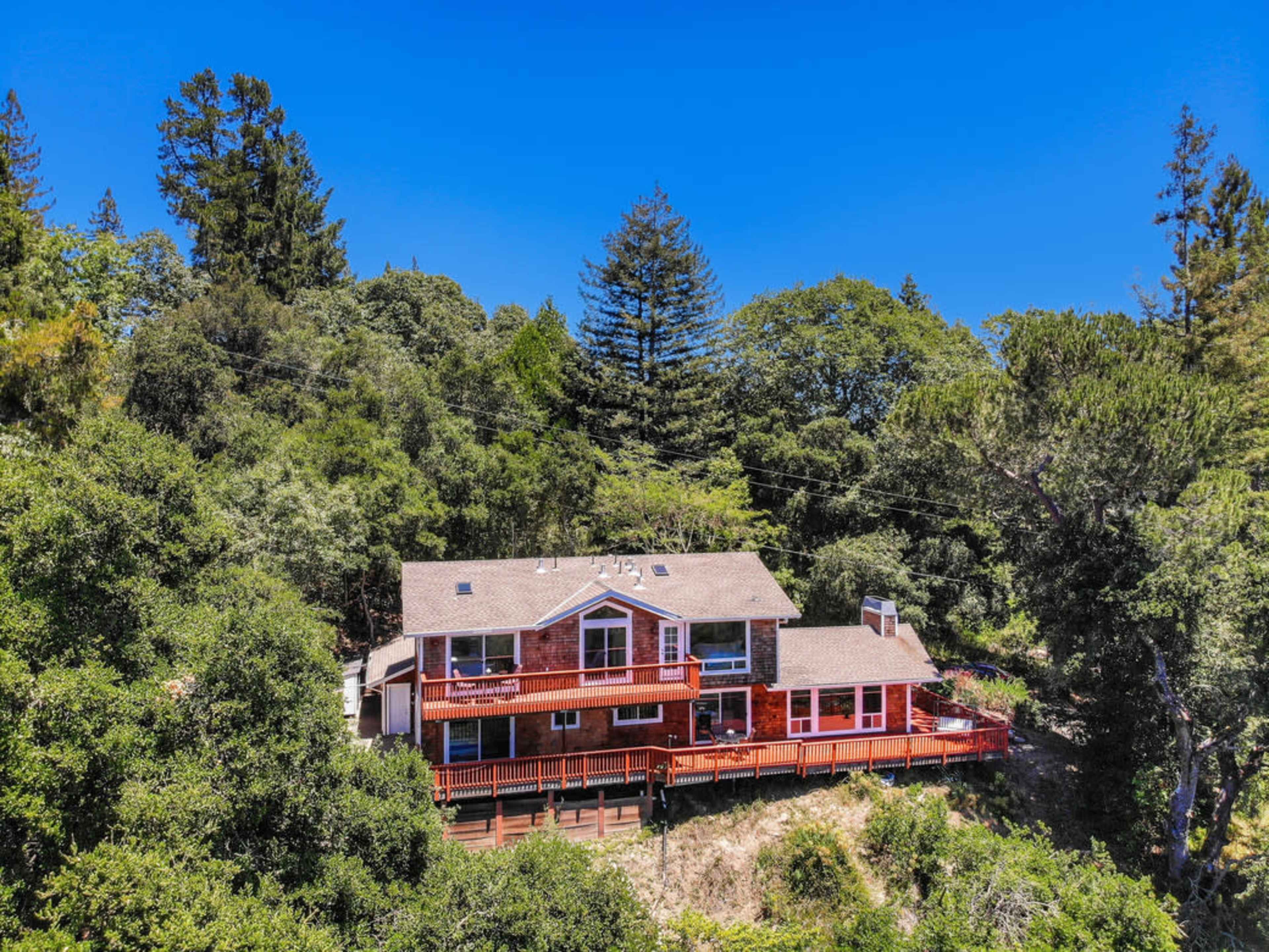 A large, red wooden house is situated among green trees on a hillside, featuring a balcony and multiple gabled roofs.