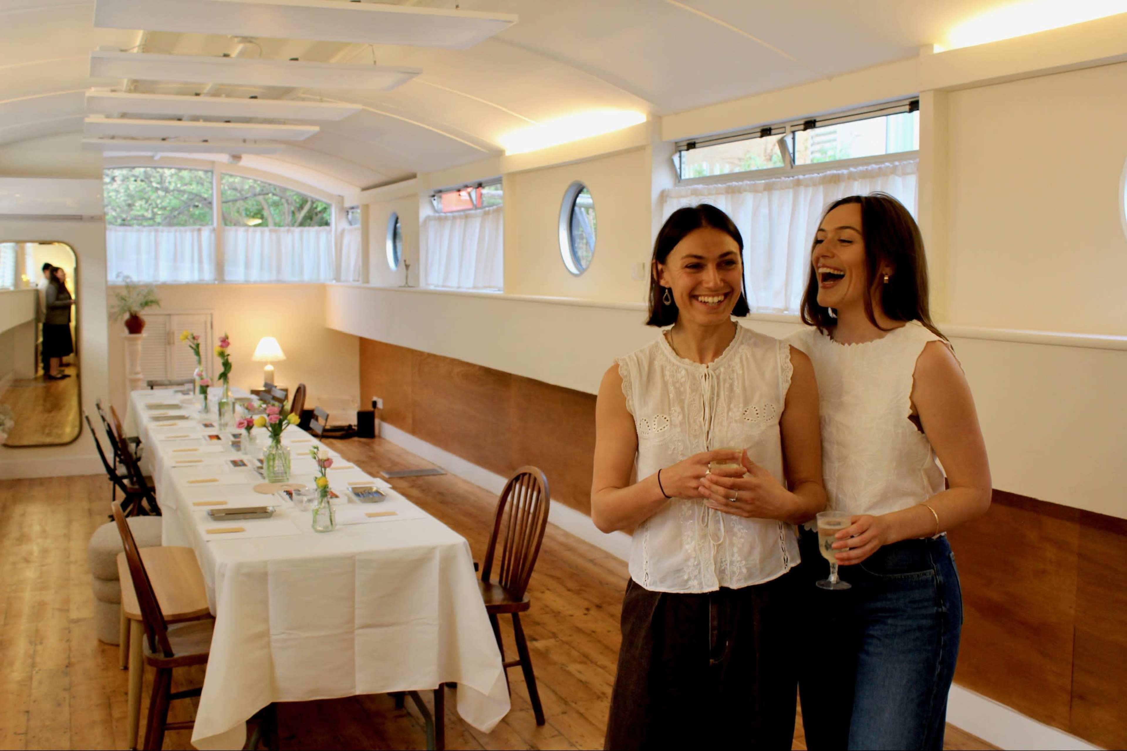 Two women stand smiling in a bright, modern dining space with a long table set for a meal.