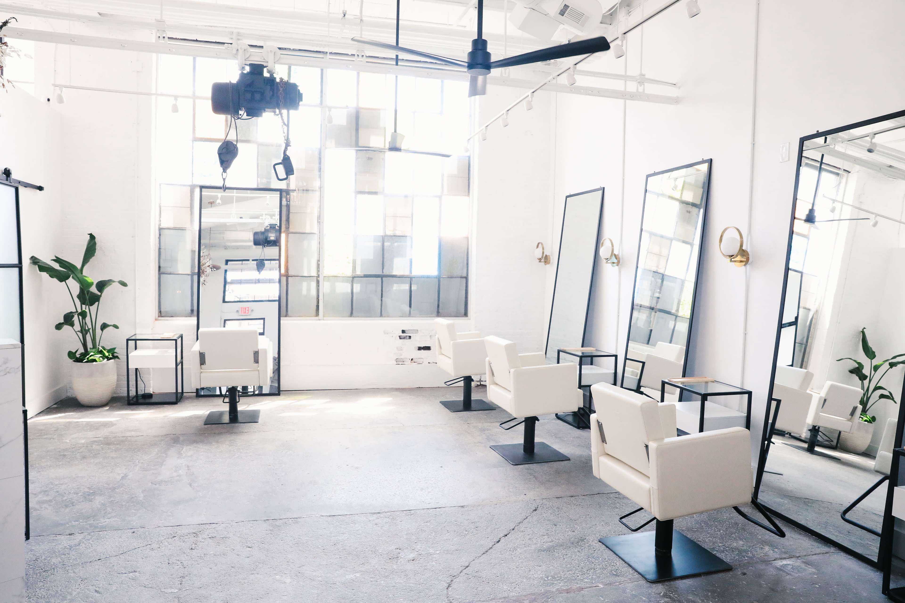 A bright, minimalist salon with white walls, large mirrors, and several hairdressing chairs arranged in a spacious layout.