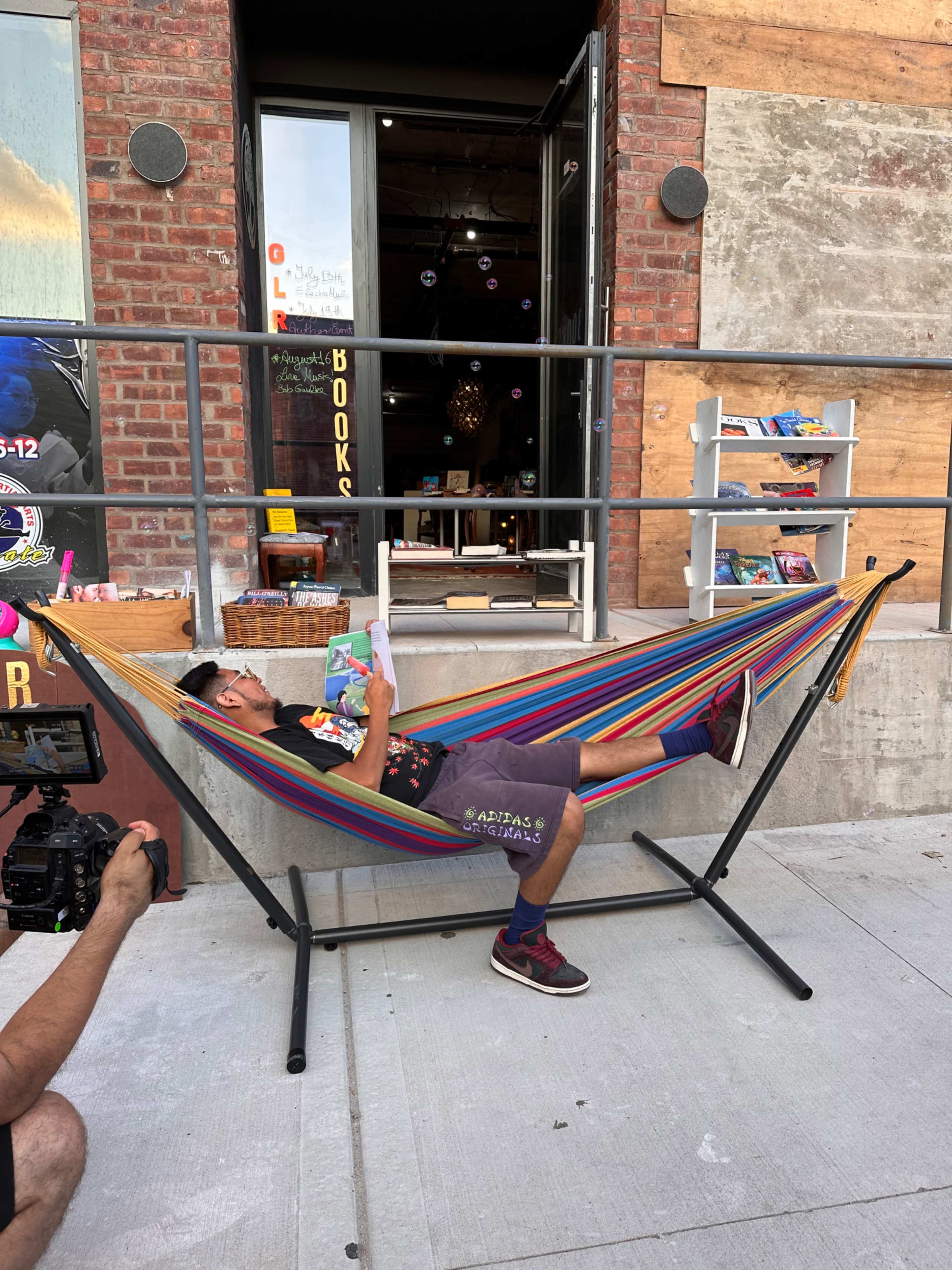 A person is lying in a colorful hammock outside a bookstore, reading a book while someone holds a camera nearby.