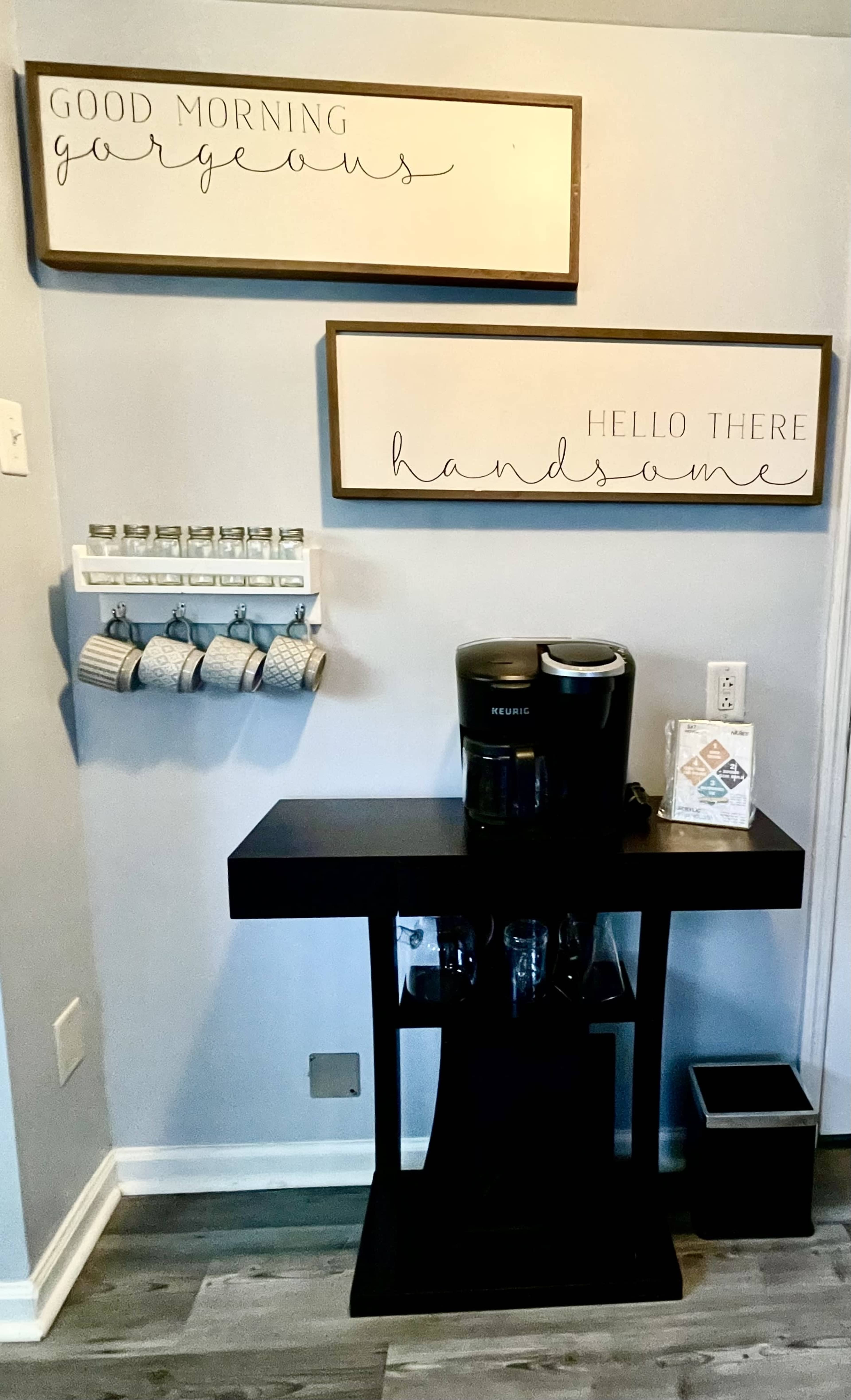 A coffee station with a Keurig machine, decorative signs on the wall, and a rack holding cups.
