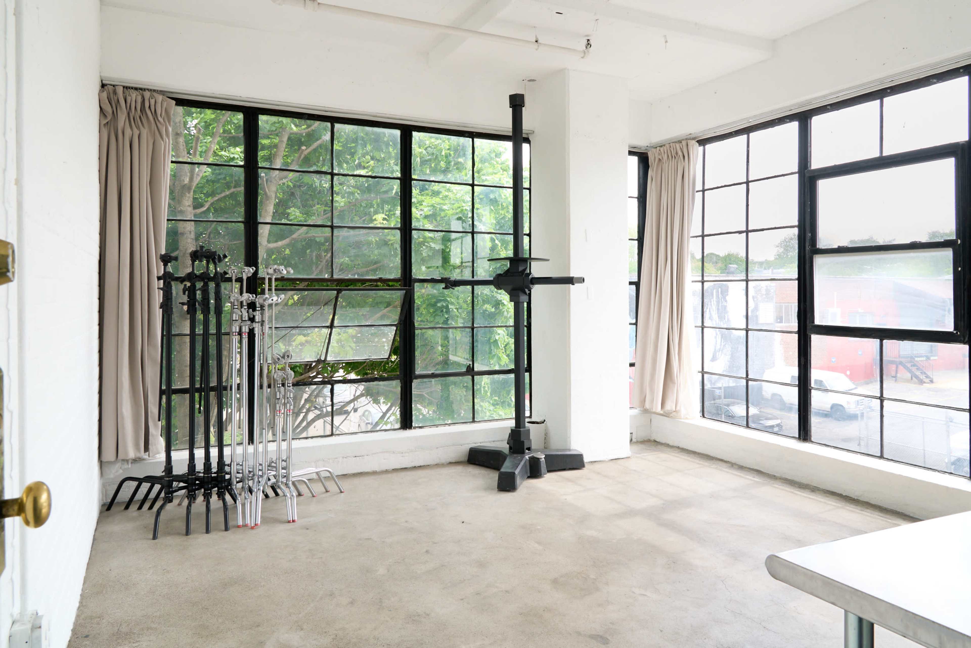 The image shows an empty room with large windows, revealing a view of greenery outside, and features multiple stands arranged along one wall.
