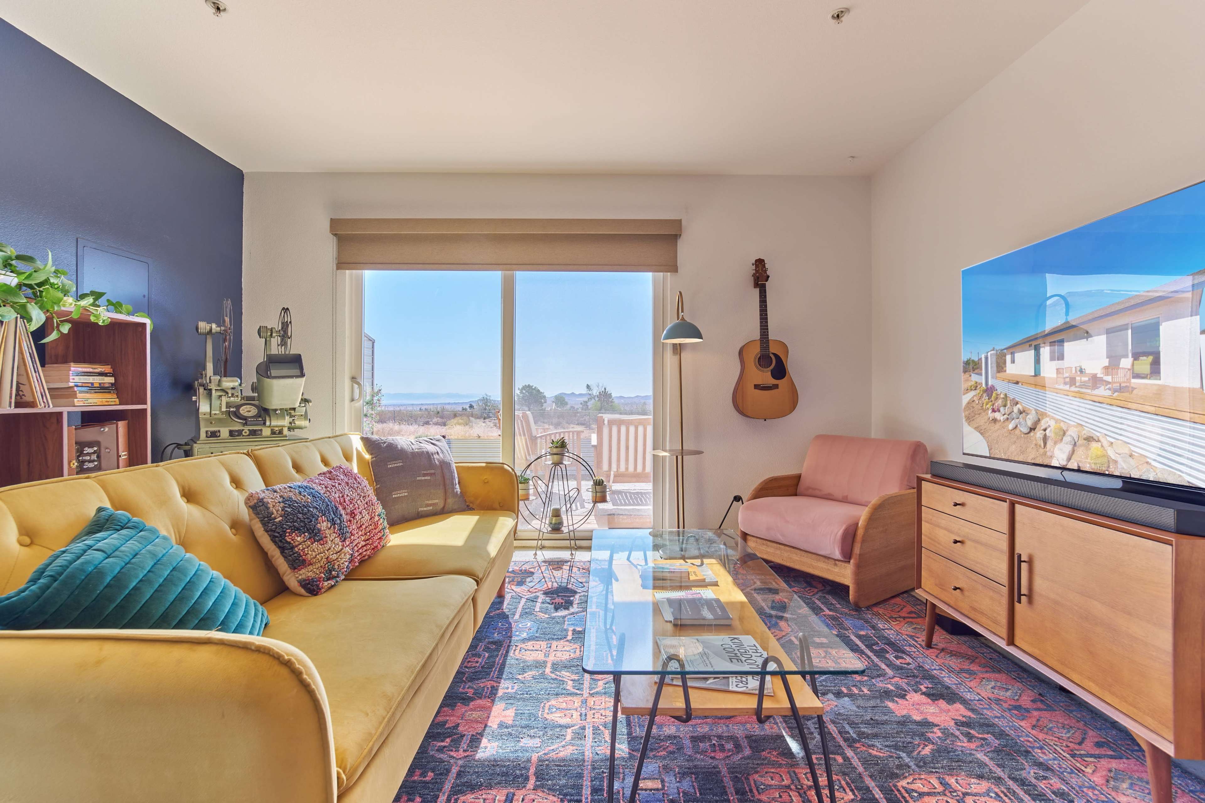 A cozy living room features a yellow sofa, a pink chair, a guitar on the wall, and a large window with a view.