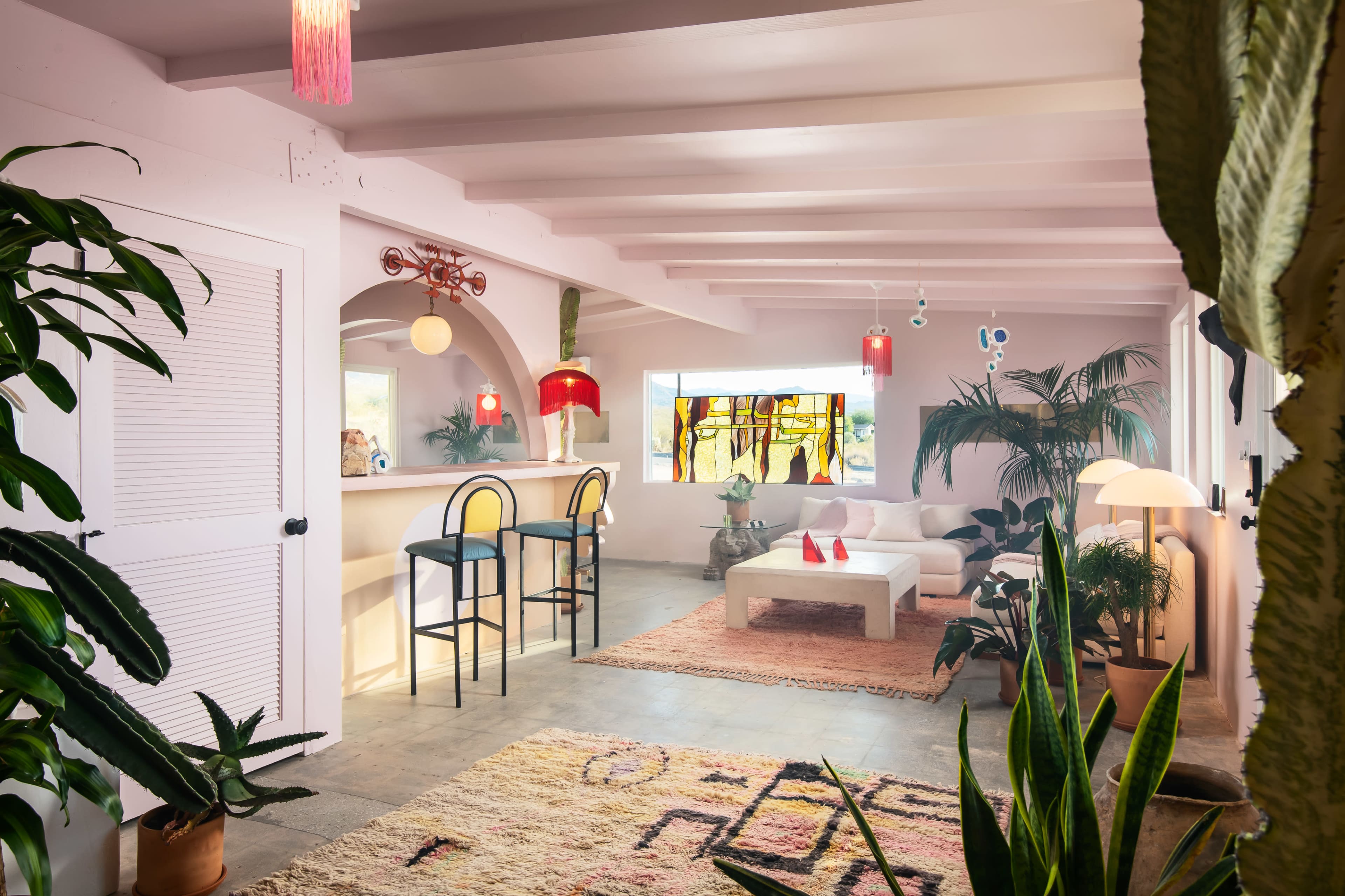 The image shows a modern living space featuring a white sofa, a bar area with two high stools, and various plants arranged throughout the room.