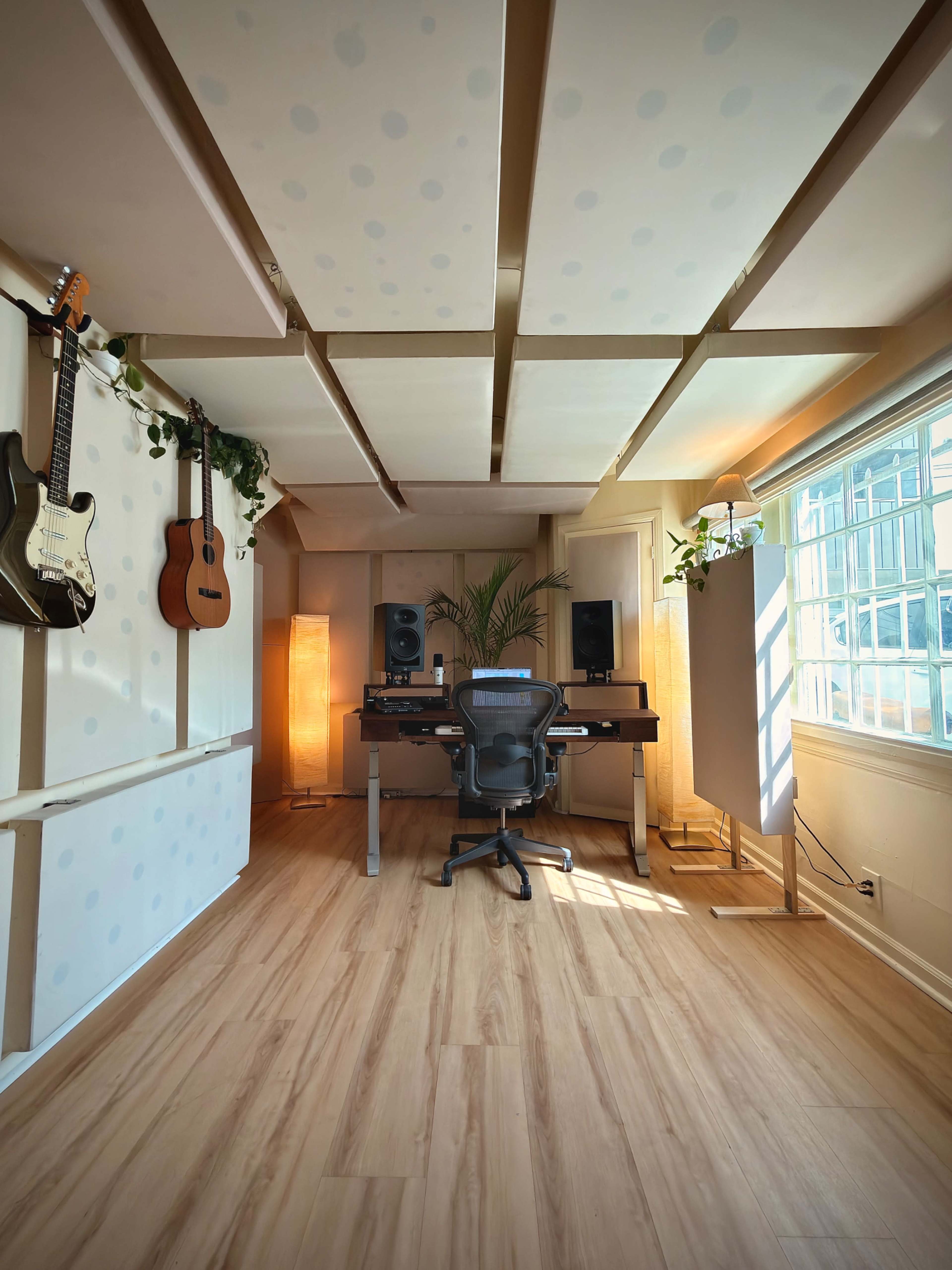 The image shows a music production studio with a chair, mixing equipment, and two guitars hanging on the wall.