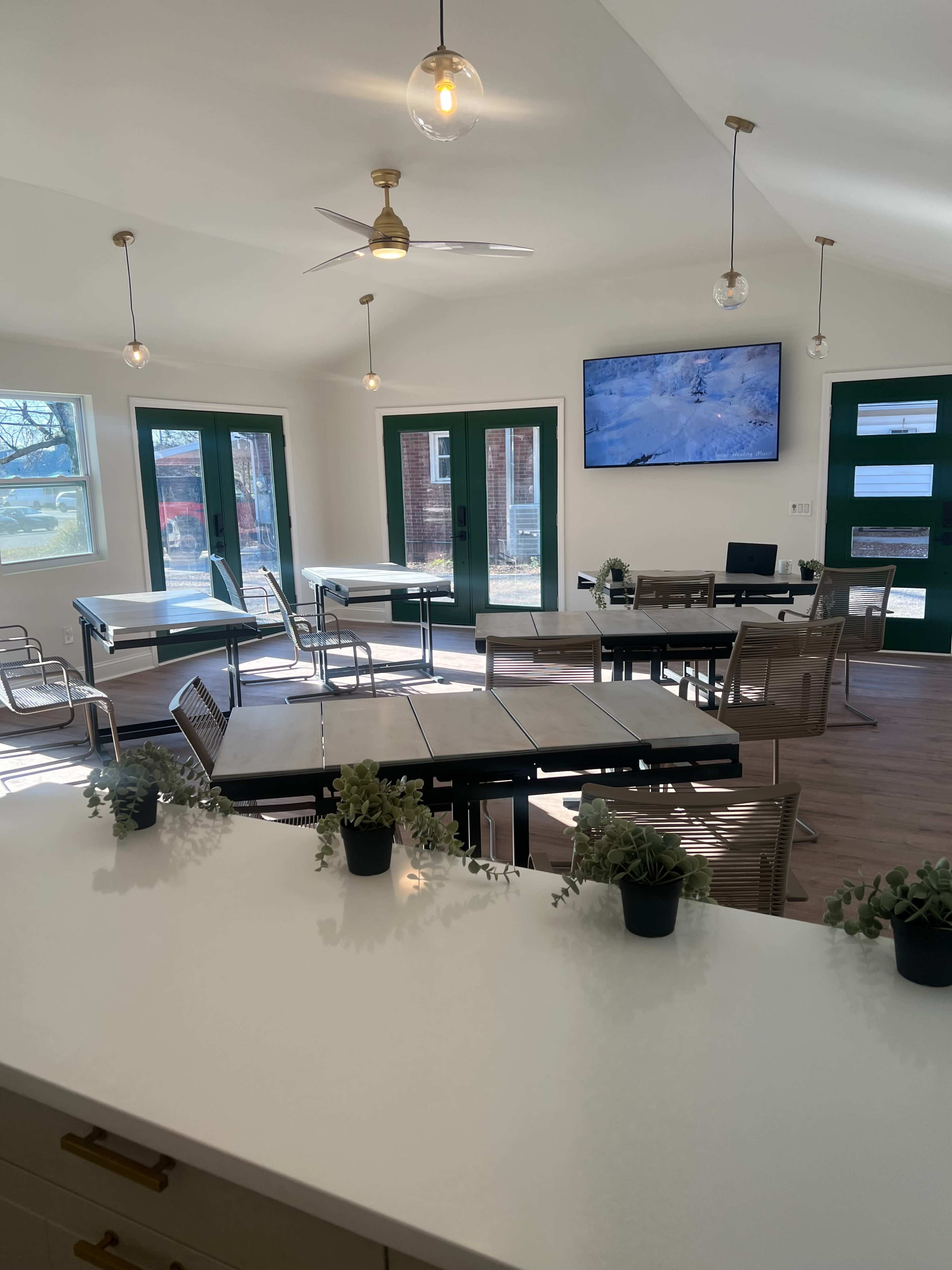 The image shows a bright, modern interior of a workspace with multiple tables and chairs, large windows, and a television mounted on the wall.