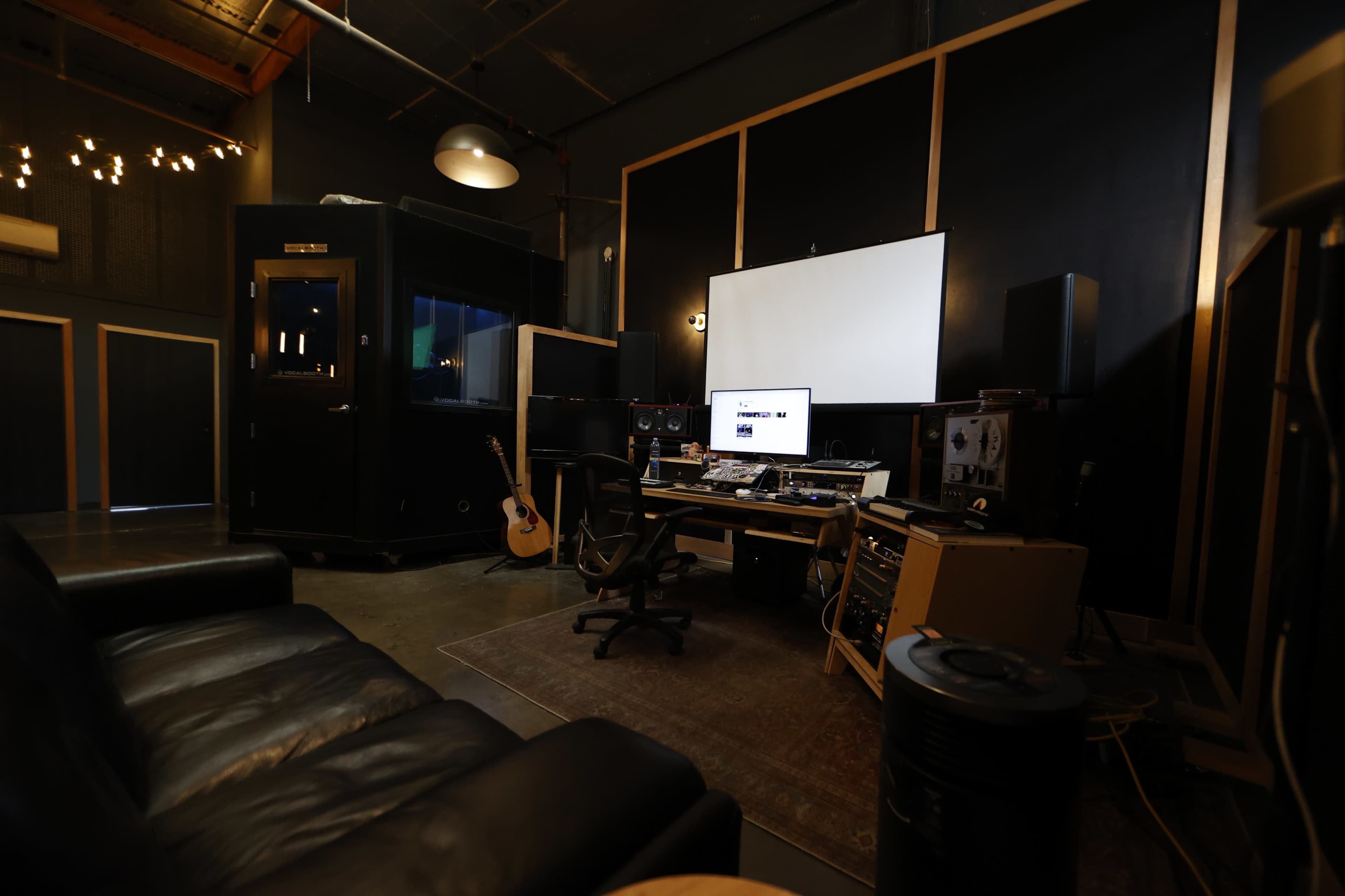 The image shows a recording studio with a desk setup, computer, audio equipment, and a guitar, surrounded by dark walls and ambient lighting.