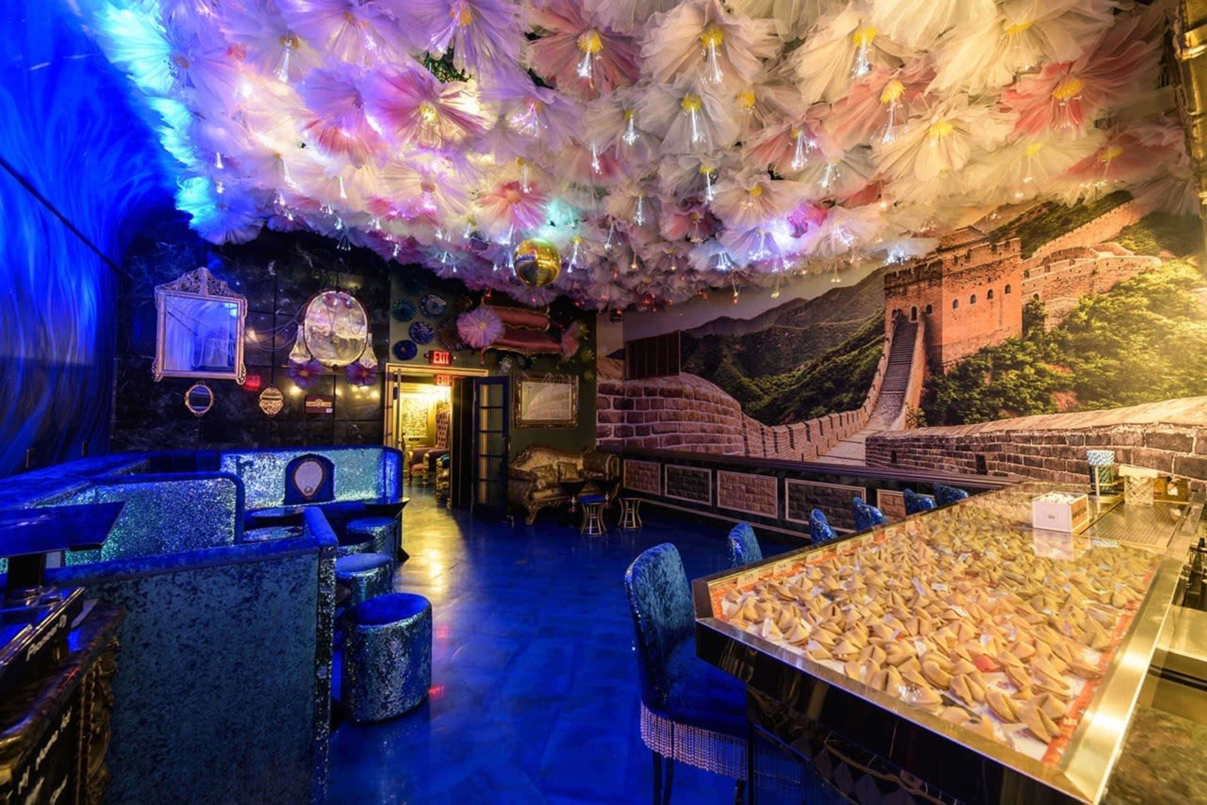 The image shows a modern bar with a shimmering metallic counter, surrounded by vibrant decor, including a ceiling adorned with colorful fabric and a mural depicting the Great Wall of China.