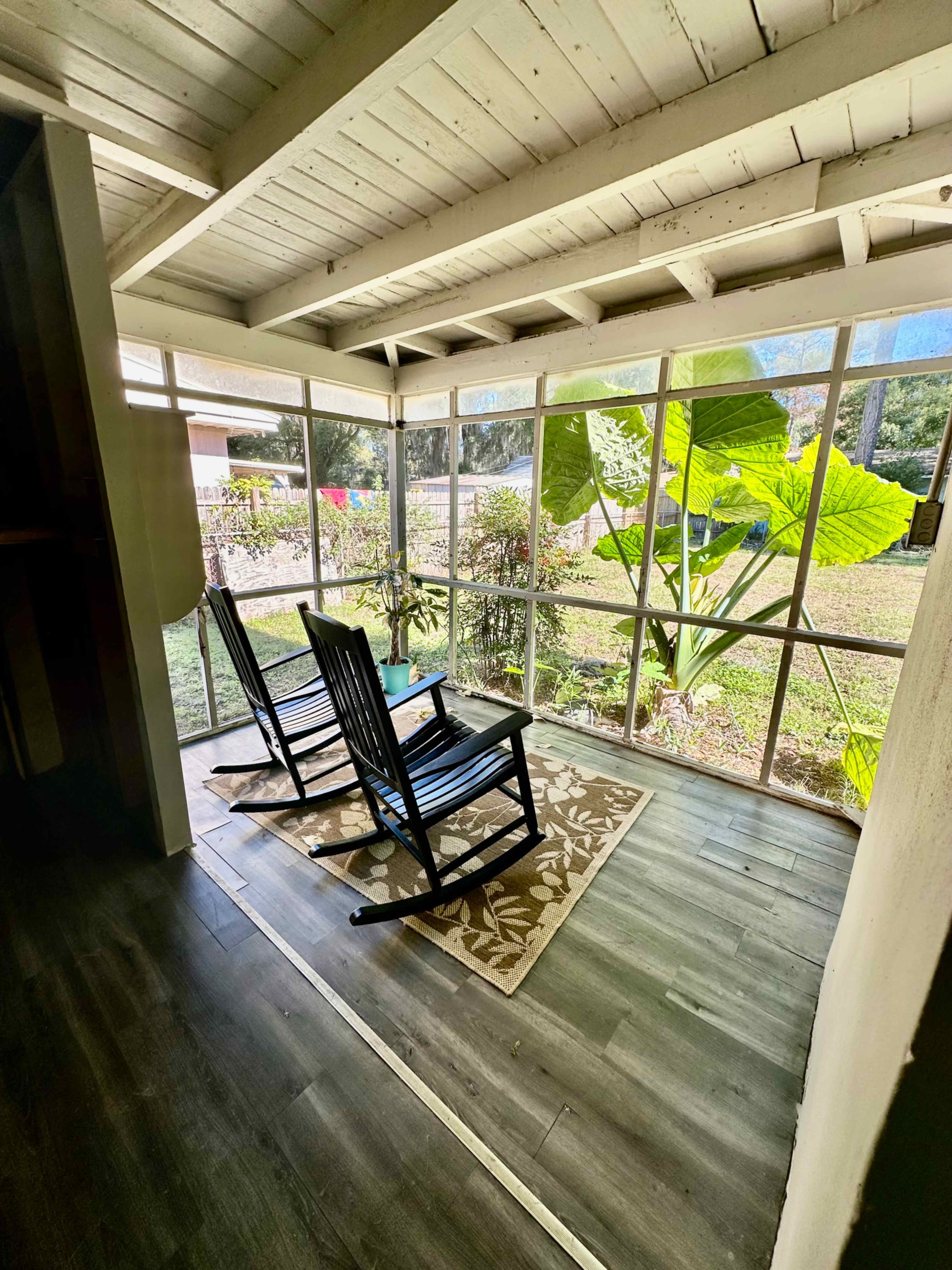 The image shows a sunlit porch with two rocking chairs facing a large window surrounded by greenery.