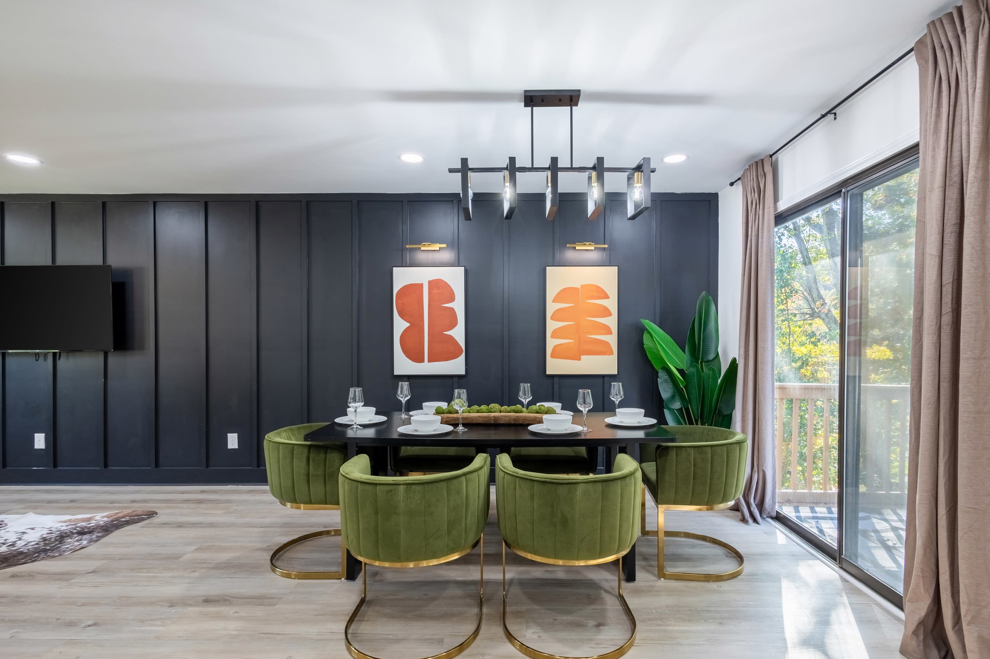 A modern dining area features a long table with green chairs, framed artwork on the dark wall, and large windows allowing natural light.