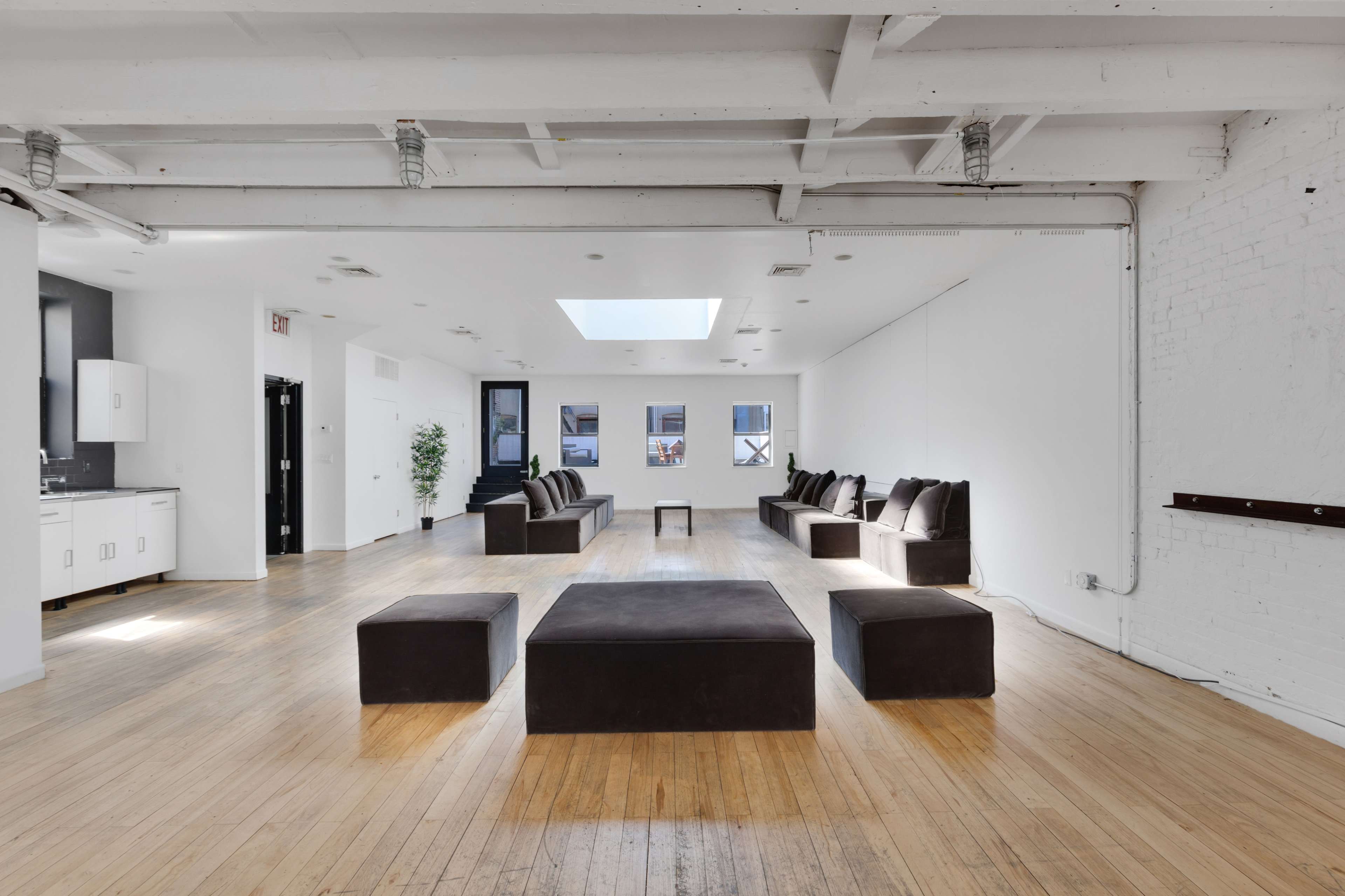 The image shows a spacious, minimalist room with wooden flooring, a large skylight, and several dark sofas arranged around a central square table.