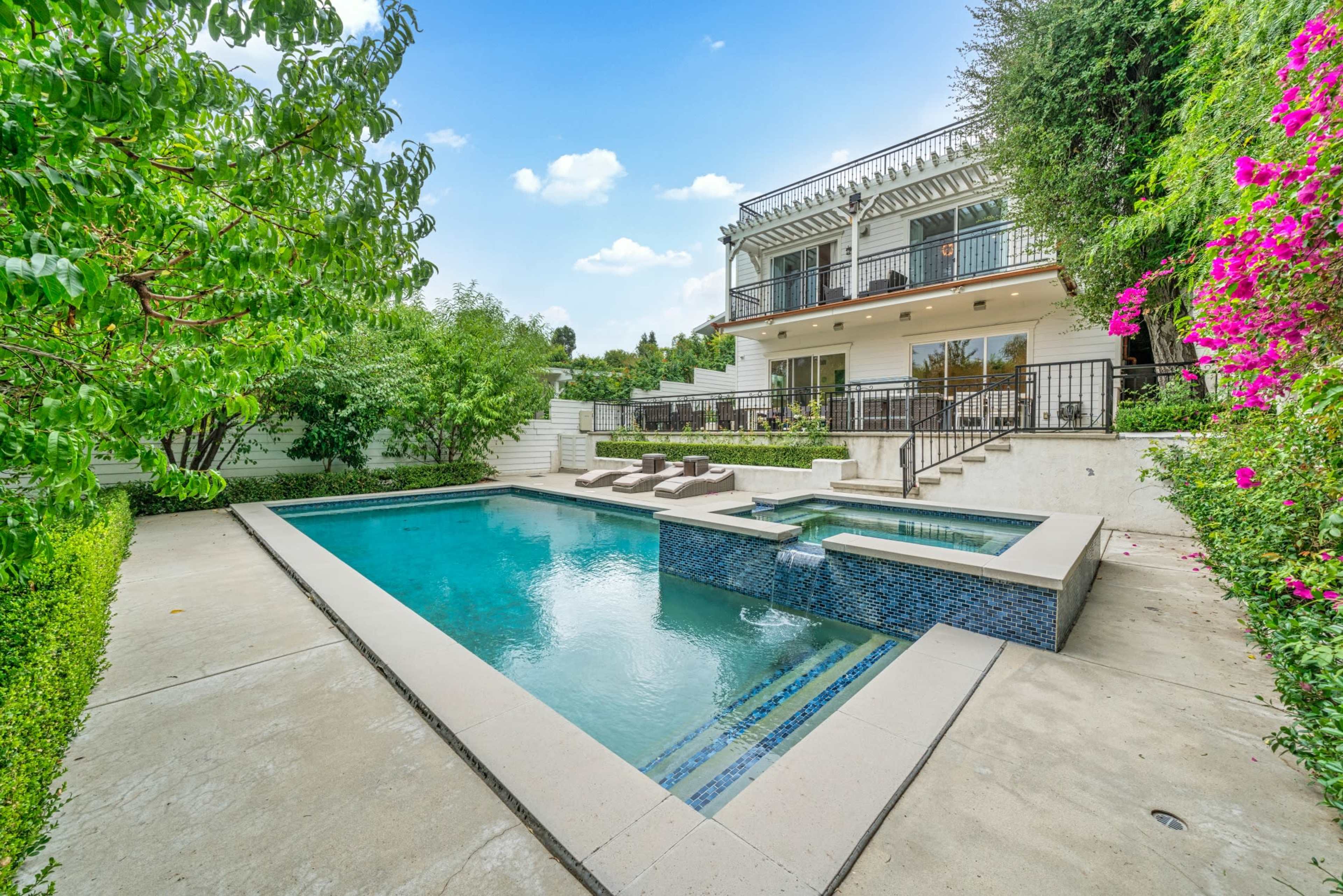 The image shows a backyard with a swimming pool and a two-story house in the background, surrounded by greenery and flowers.