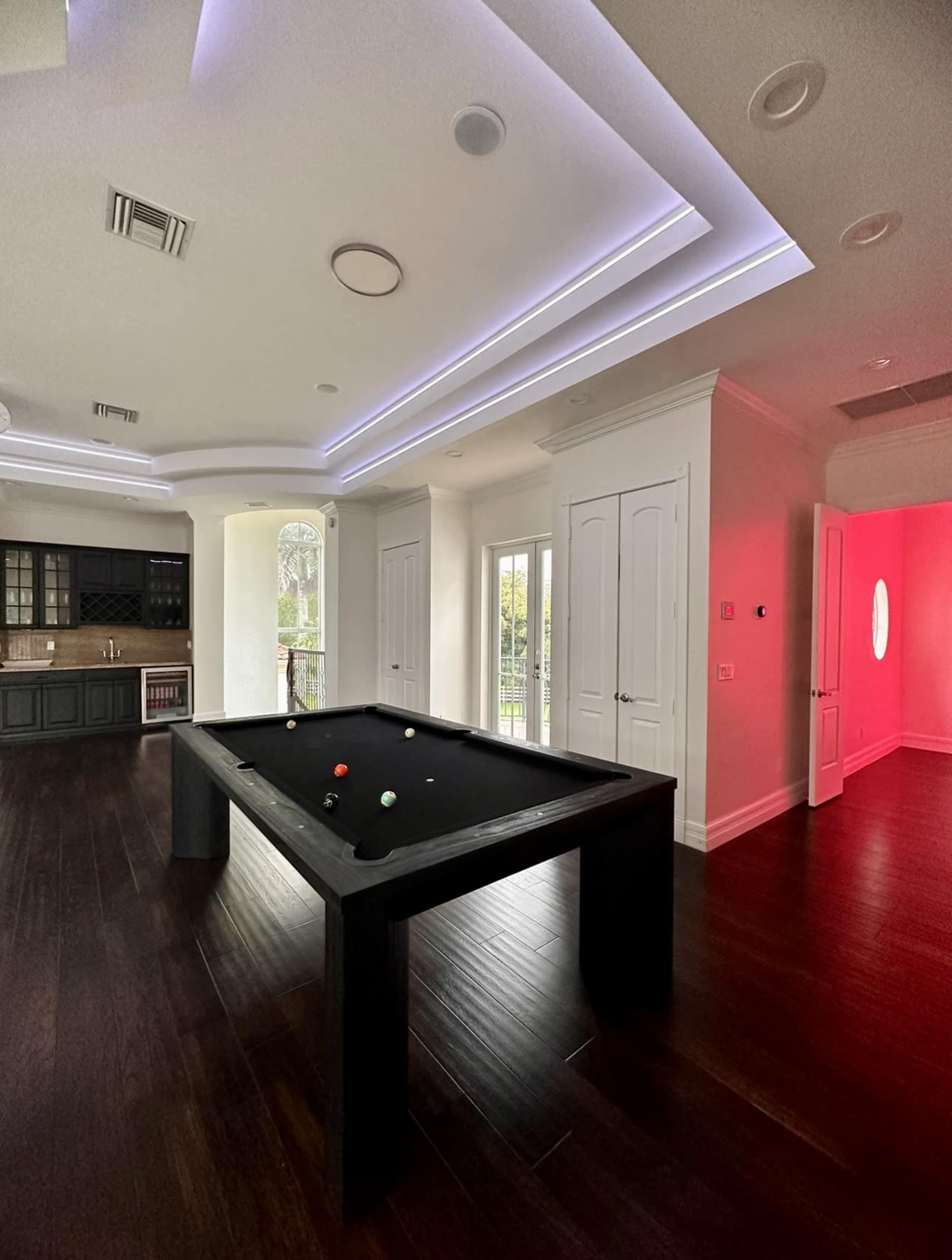 A pool table is positioned in a room with modern lighting and large windows, adjacent to a kitchen area and a corridor with a red-lit space.
