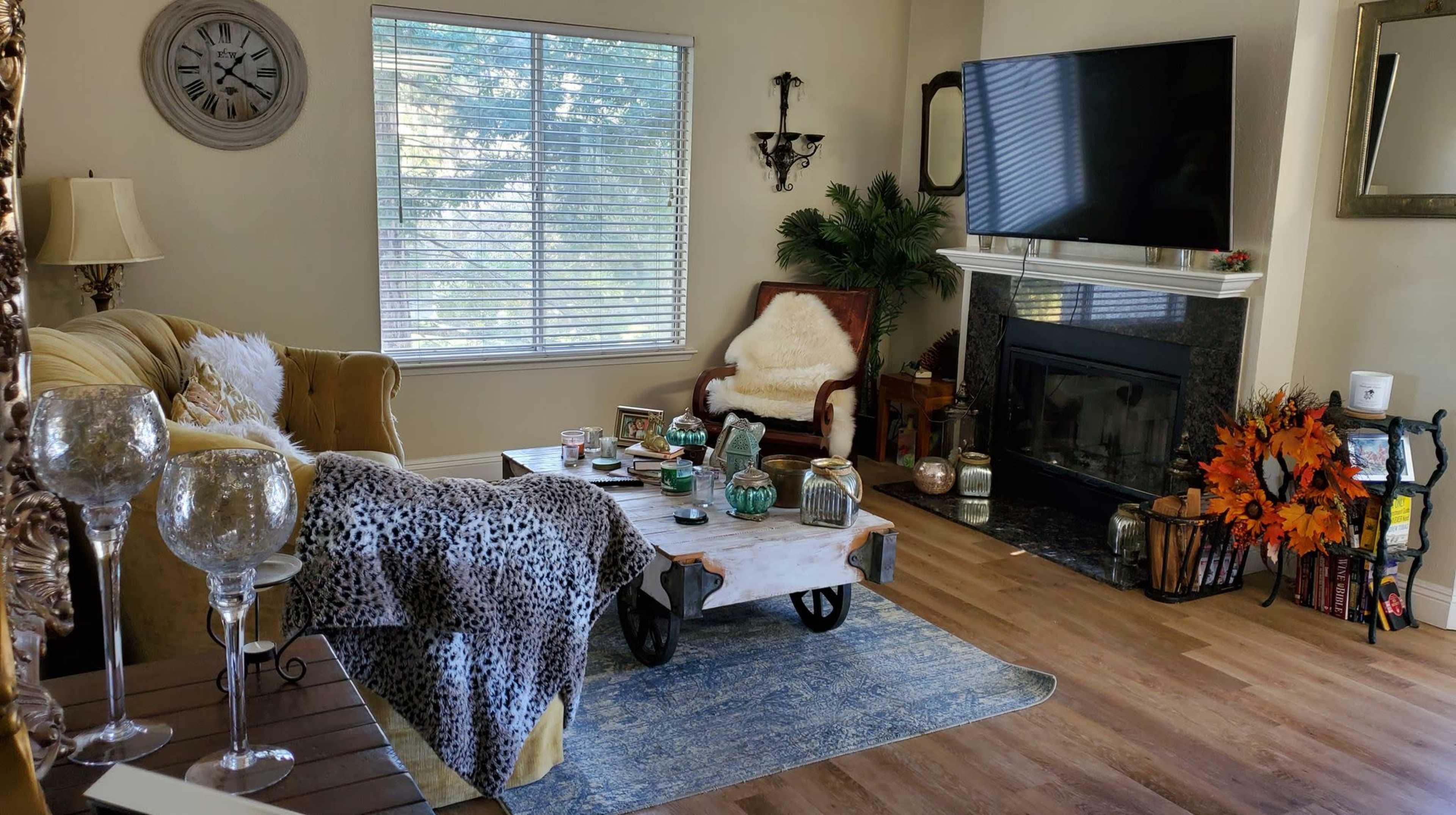 The living room features a cozy arrangement with a yellow sofa, a wooden coffee table, and a large television above a fireplace, all illuminated by natural light from a window.