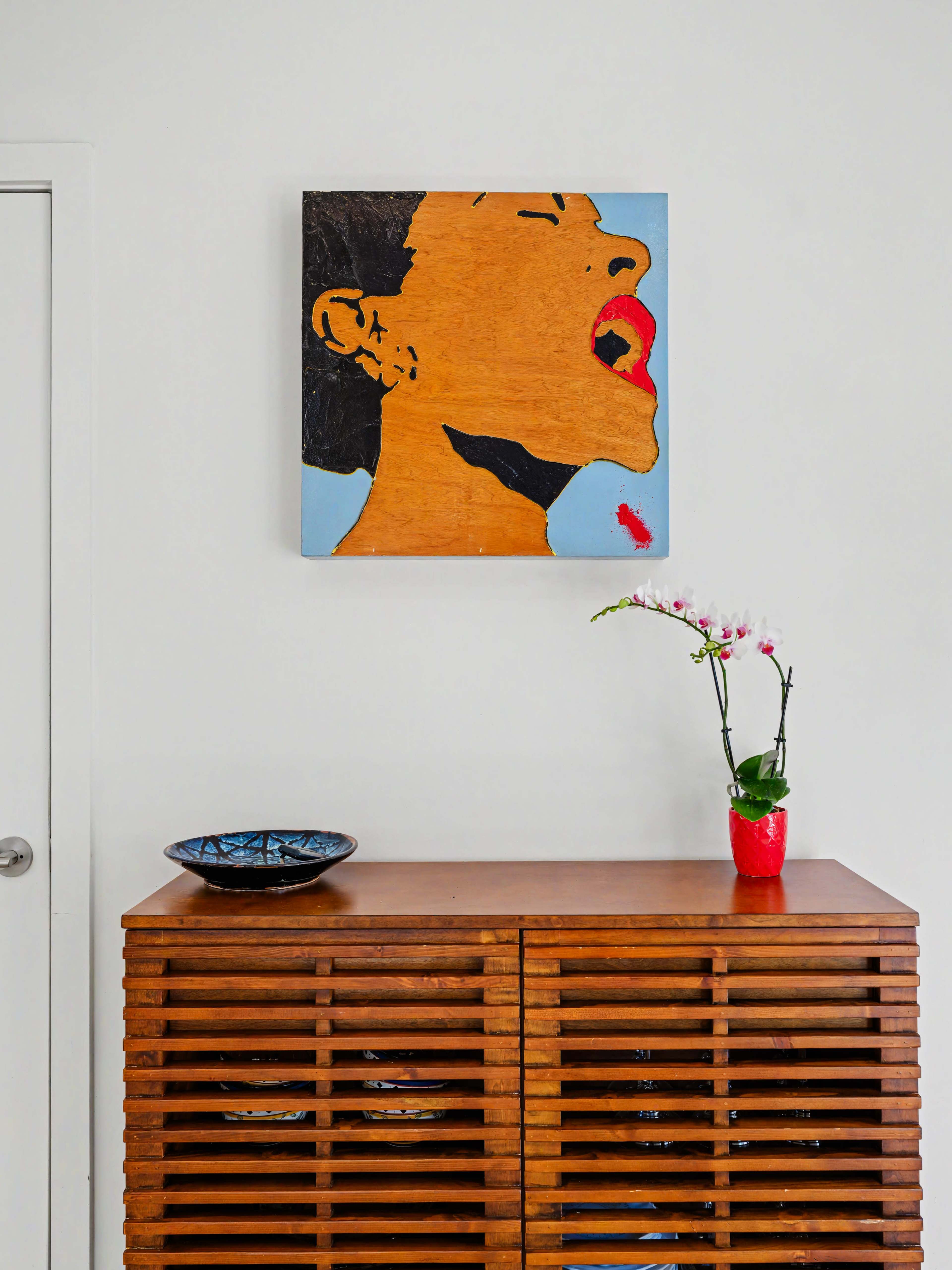 A wooden sideboard with slatted doors displays a decorative bowl and a potted orchid, with a framed artwork of a person's face in an expressive pose hanging above it.