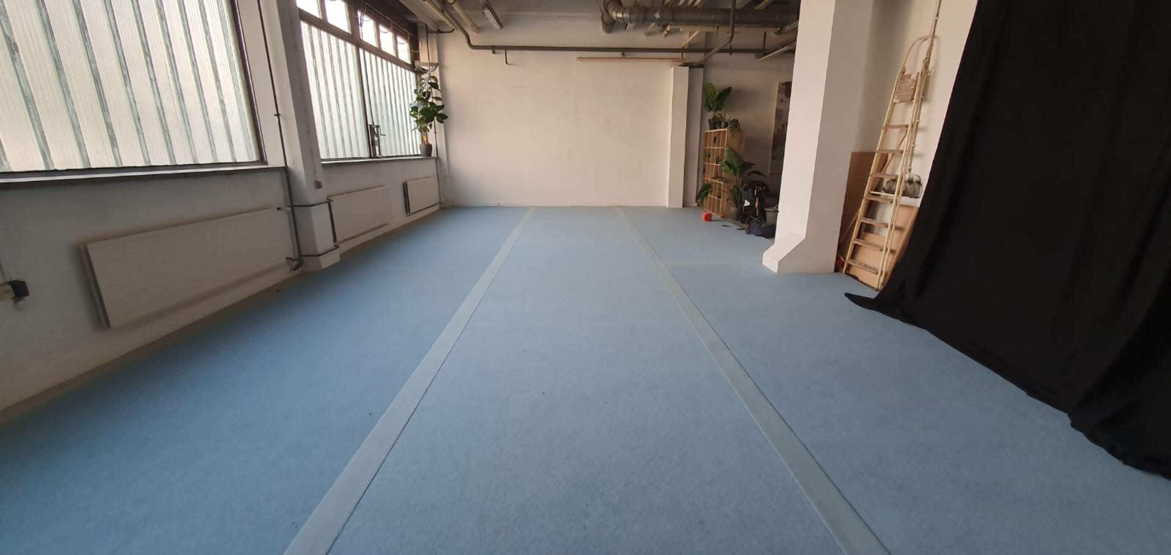 A spacious, empty room with blue carpet, large windows allowing light in, and a black curtain on one side.