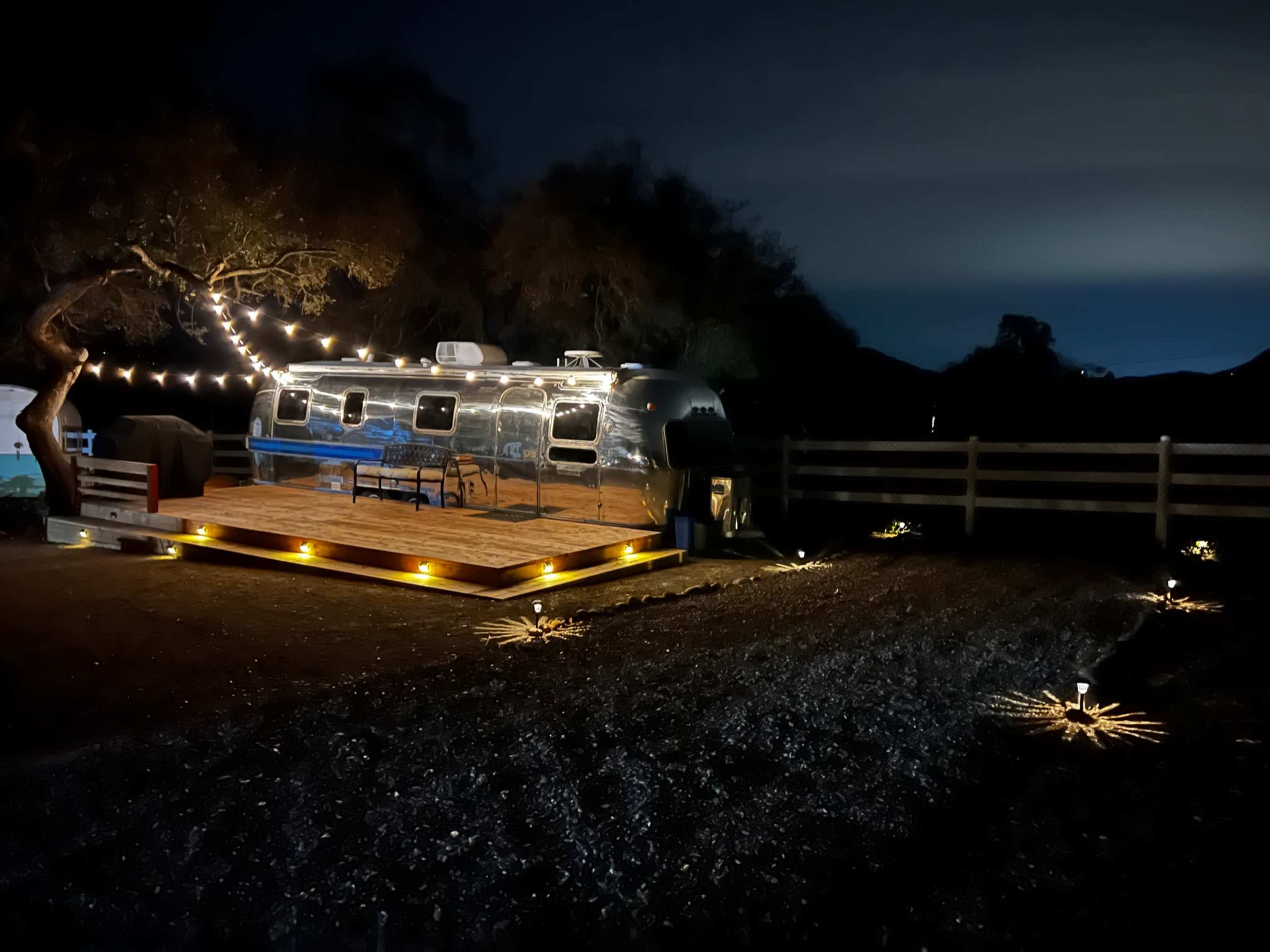 An Airstream trailer is parked on a wooden deck illuminated by string lights in a dark outdoor setting.