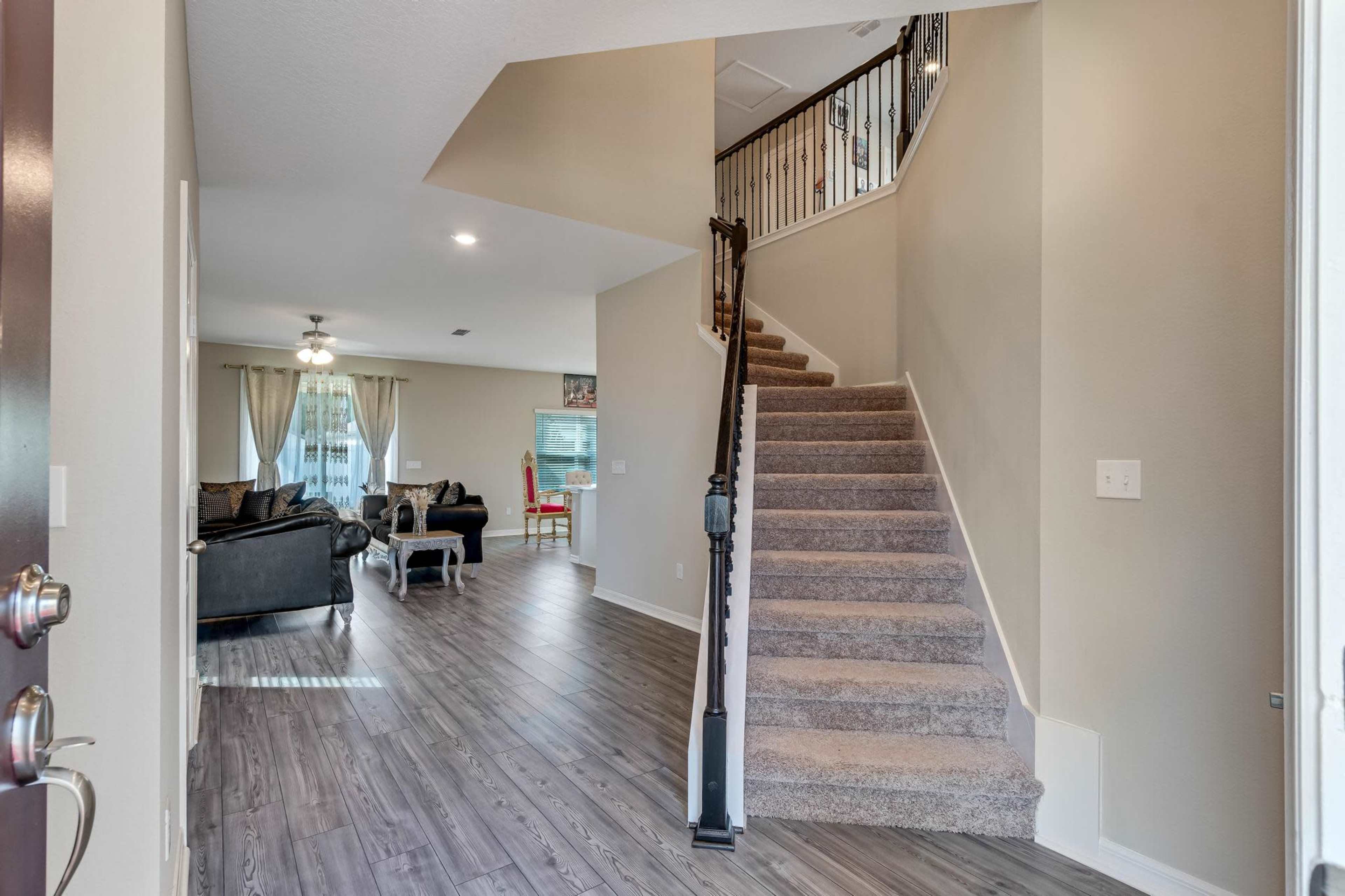 A foyer leading to a stairway, with an open living area visible in the background furnished with a couch and chairs.