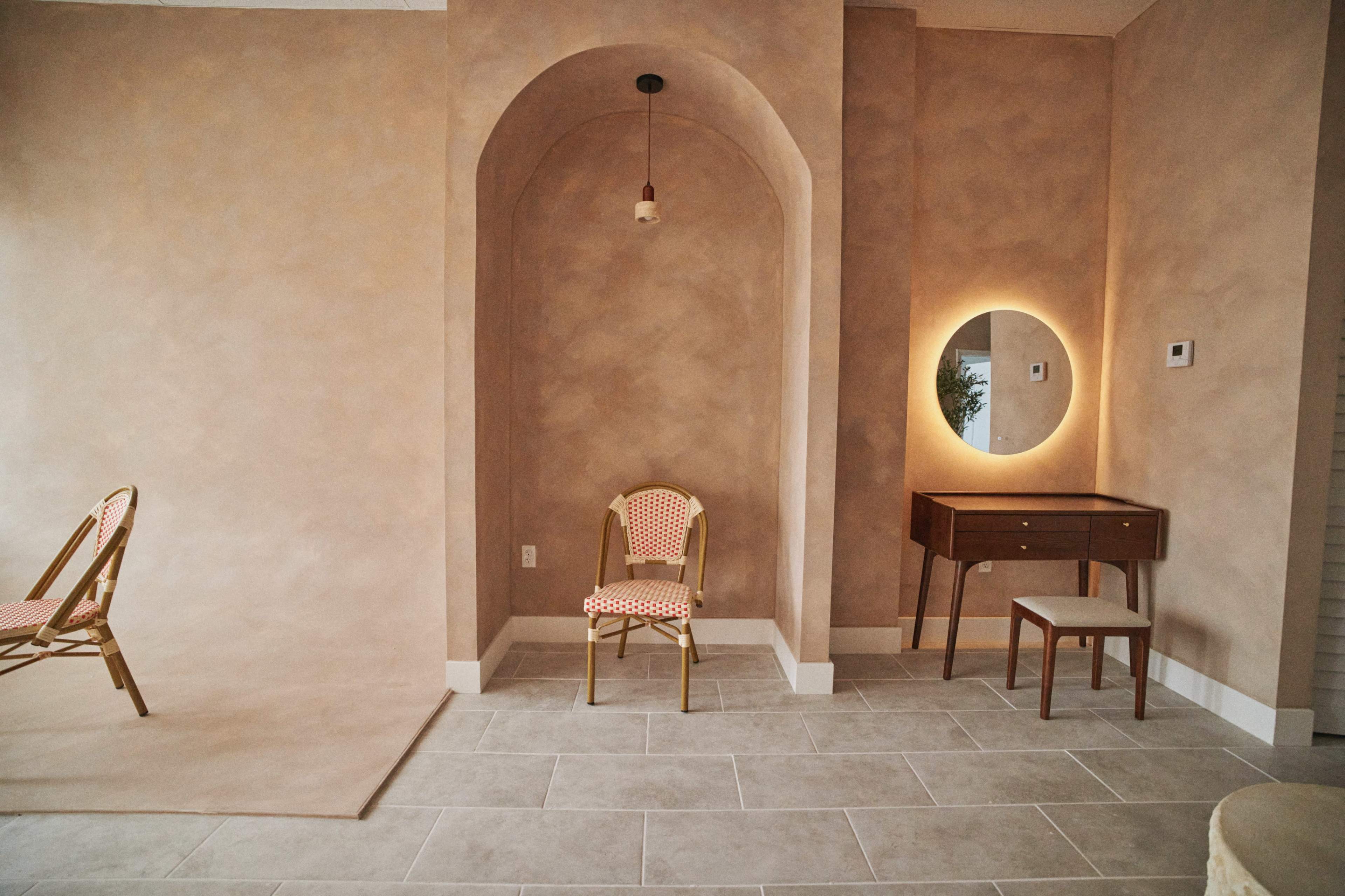 The image depicts a softly lit room with a textured beige wall, featuring a round mirror, a wooden console table, a small stool, and a patterned chair.