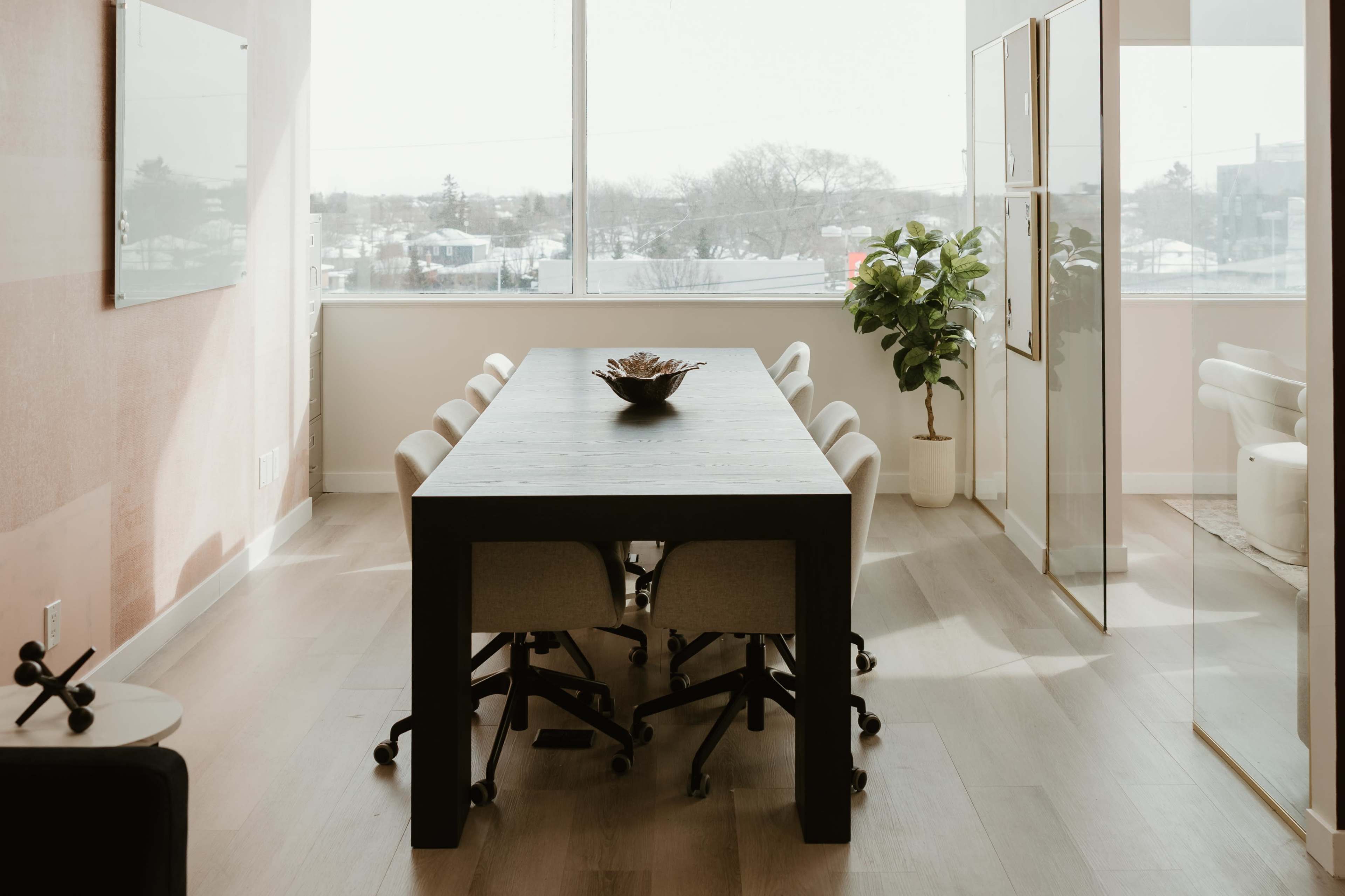 A large, rectangular wooden table with eight chairs sits in a bright, modern conference room featuring large windows and a potted plant.