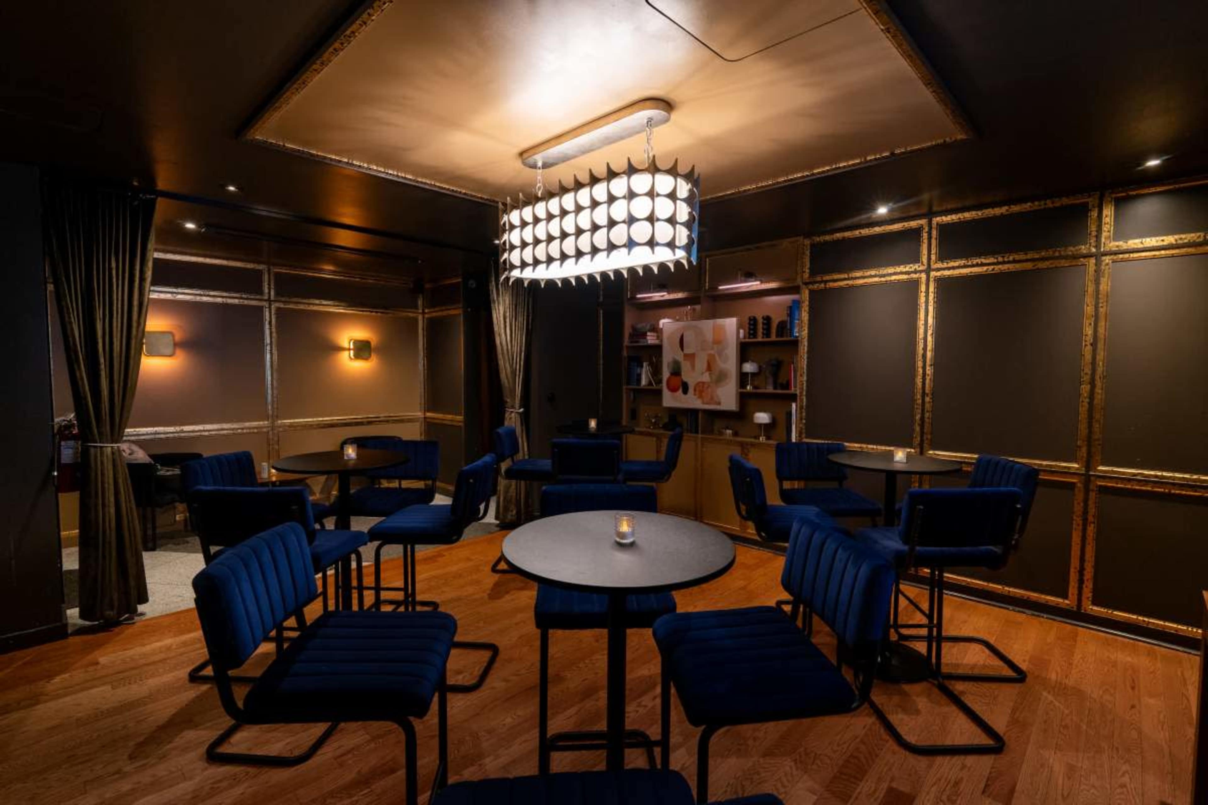 The image depicts a modern, dimly lit lounge area featuring several small tables and blue upholstered chairs arranged around a central chandelier.