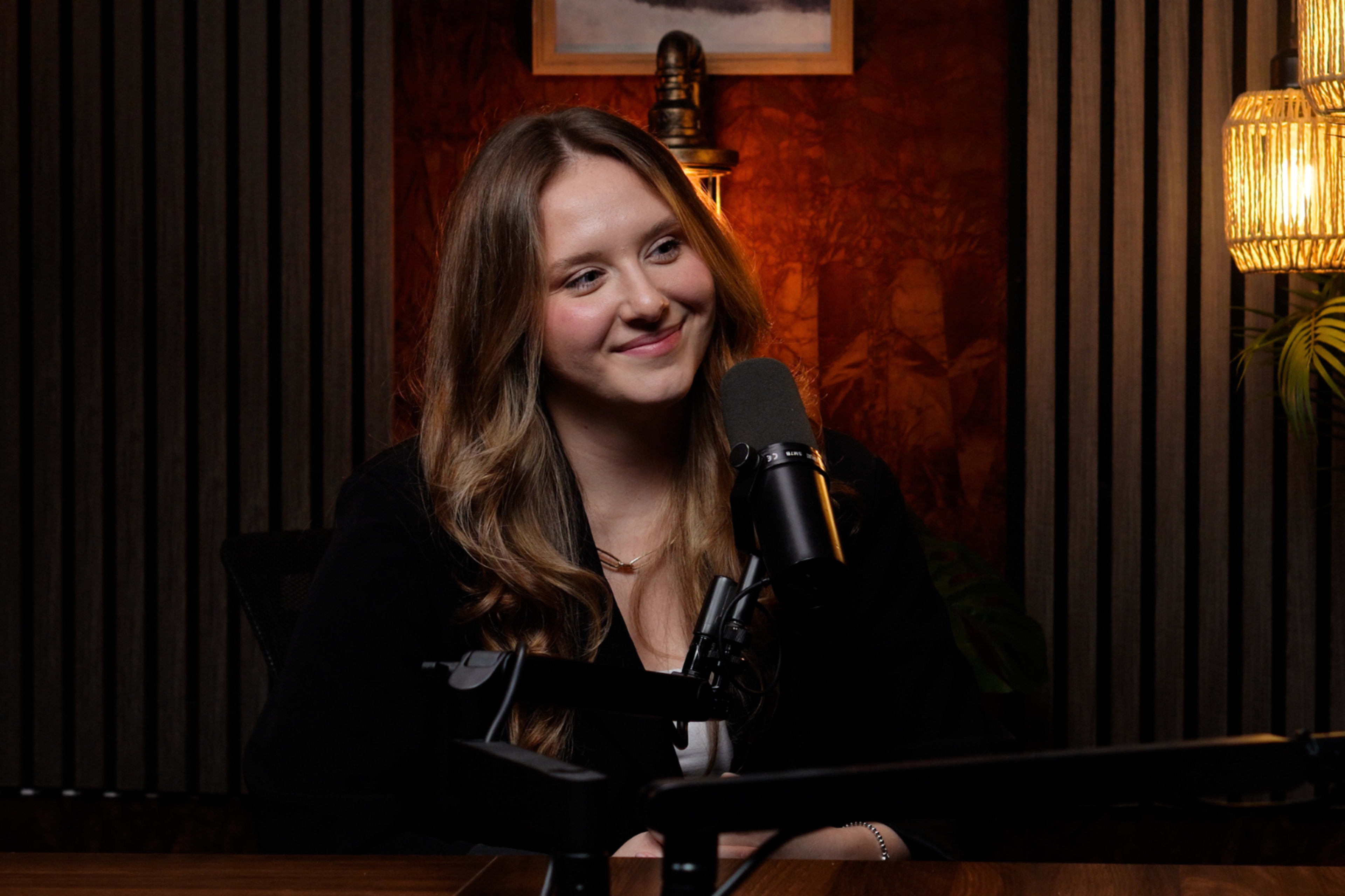 A young woman with long hair sits at a microphone in a well-lit studio with wooden accents and decorative plants.