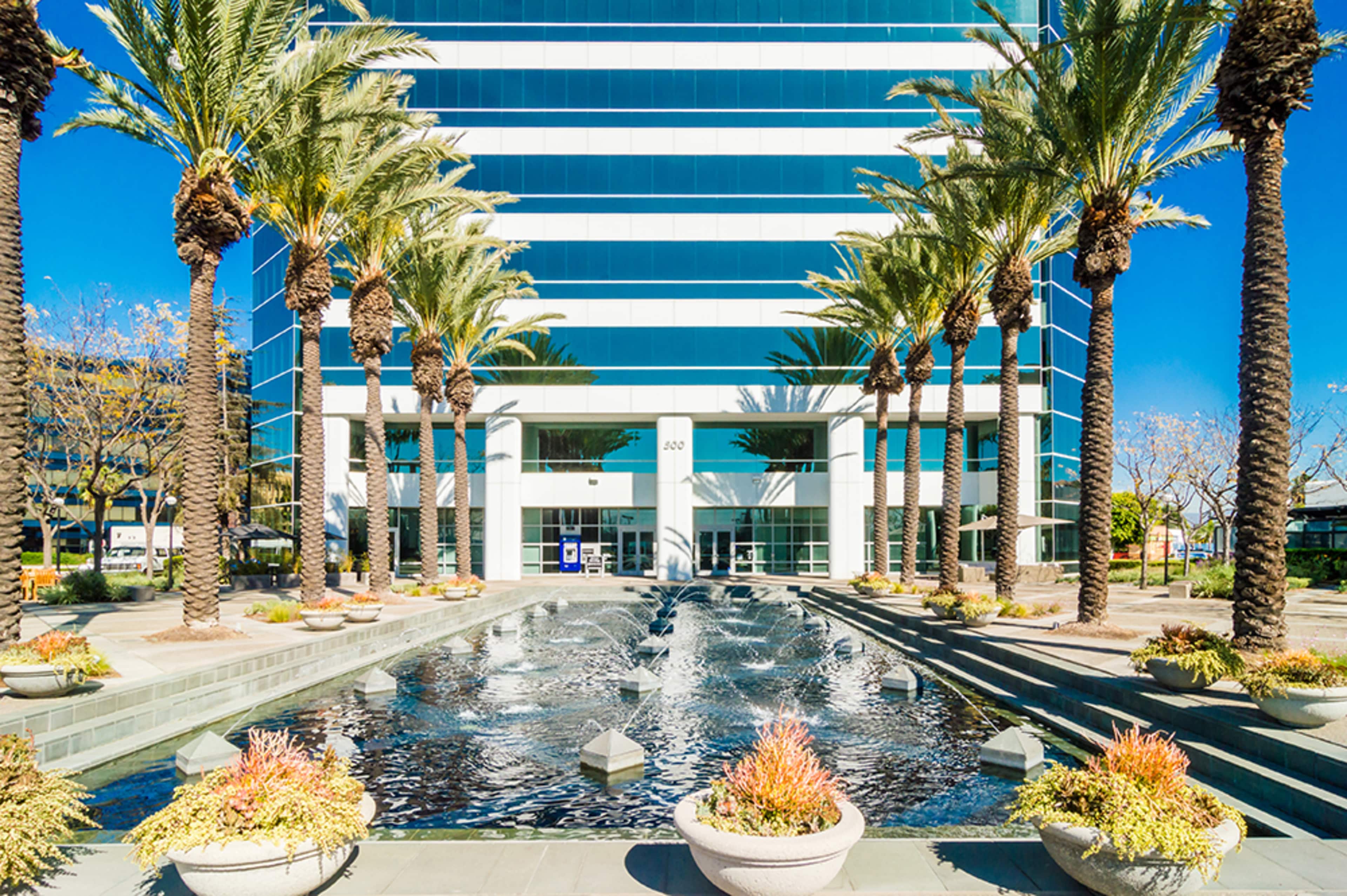 A modern office building is flanked by palm trees and features a water fountain in the foreground.