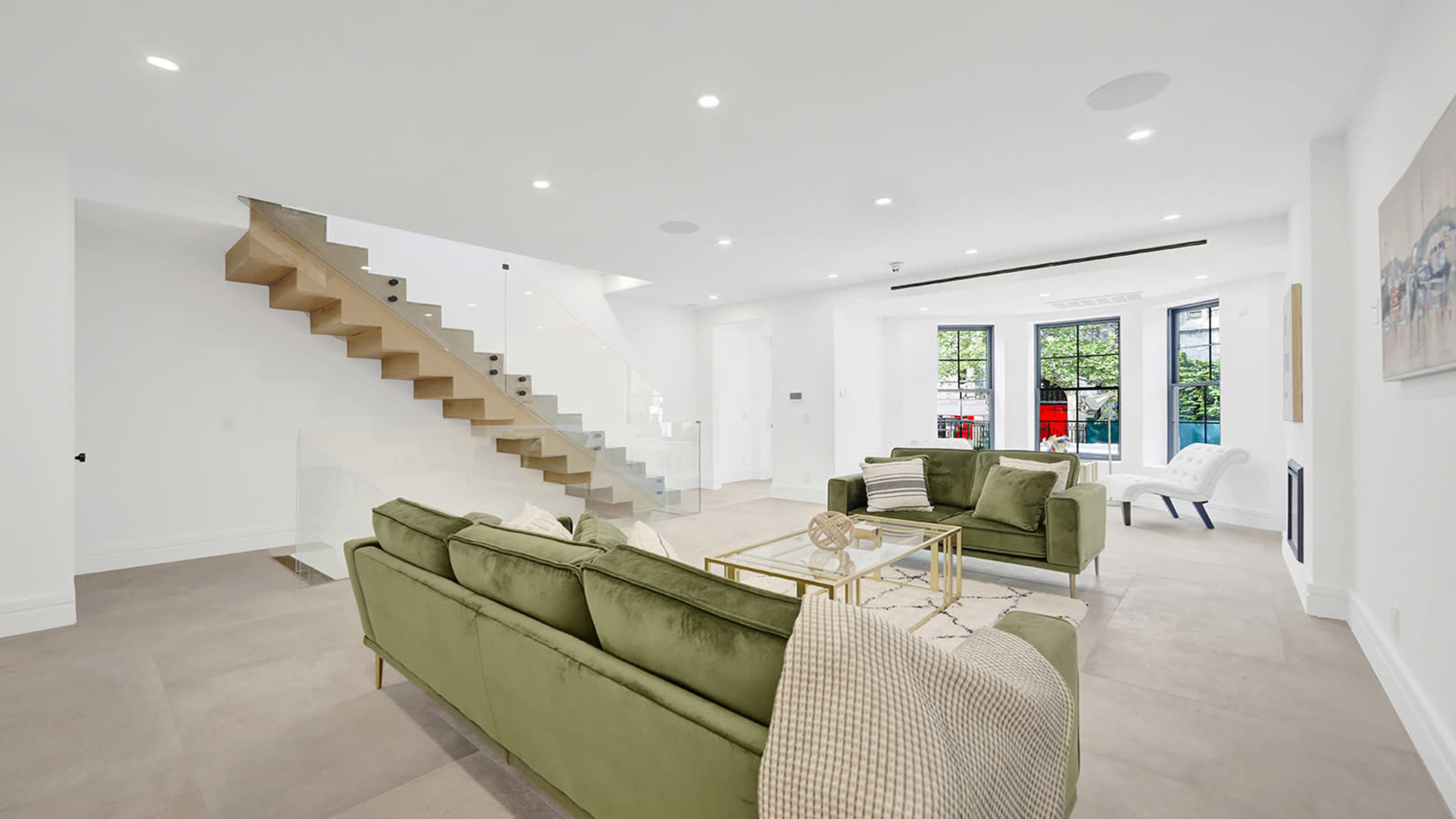 A modern living room with green sofas, a glass coffee table, and a staircase leading to the upper floor.