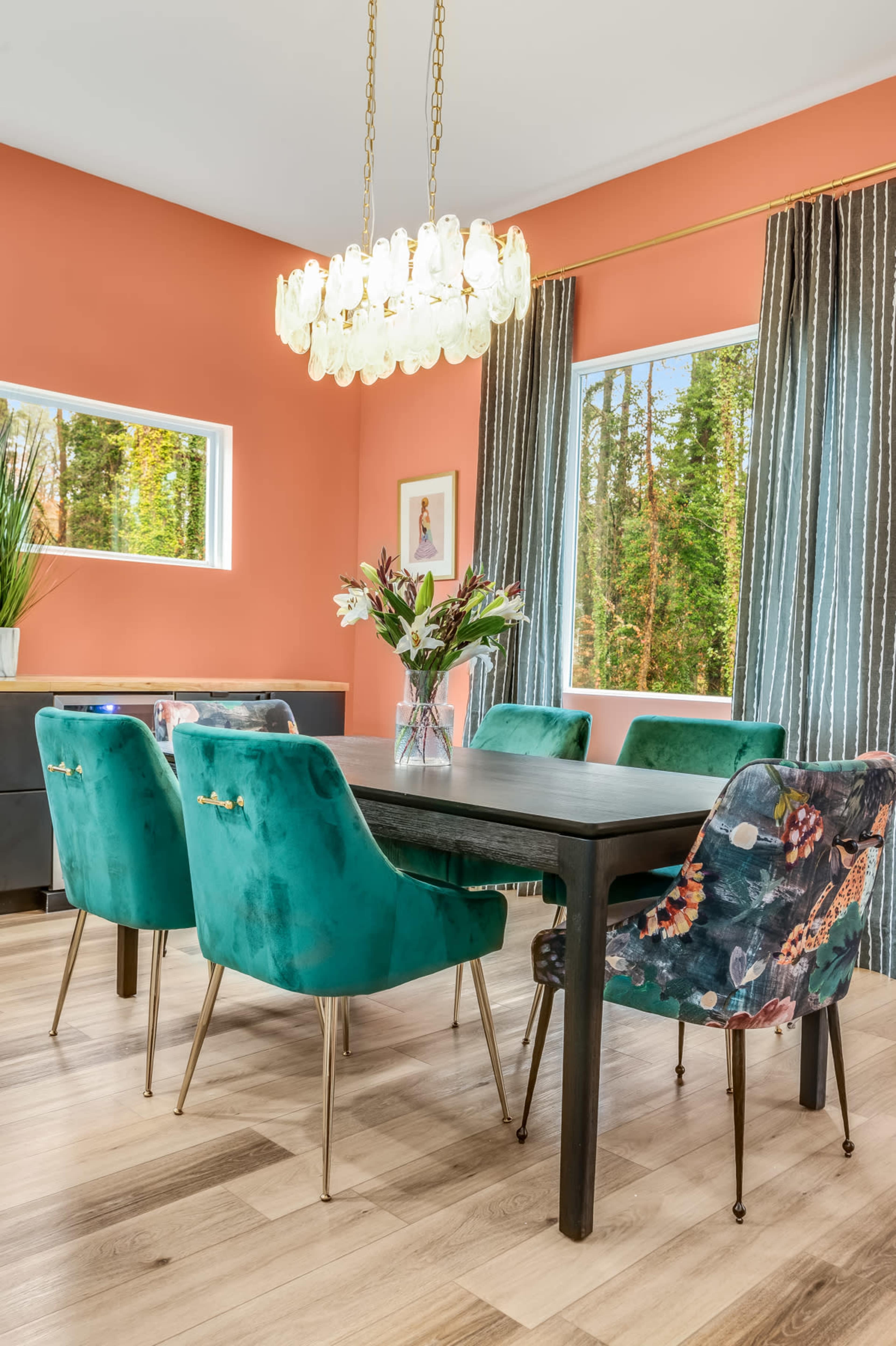 A dining room features a black table surrounded by green and patterned chairs, illuminated by a crystal chandelier against a coral wall.